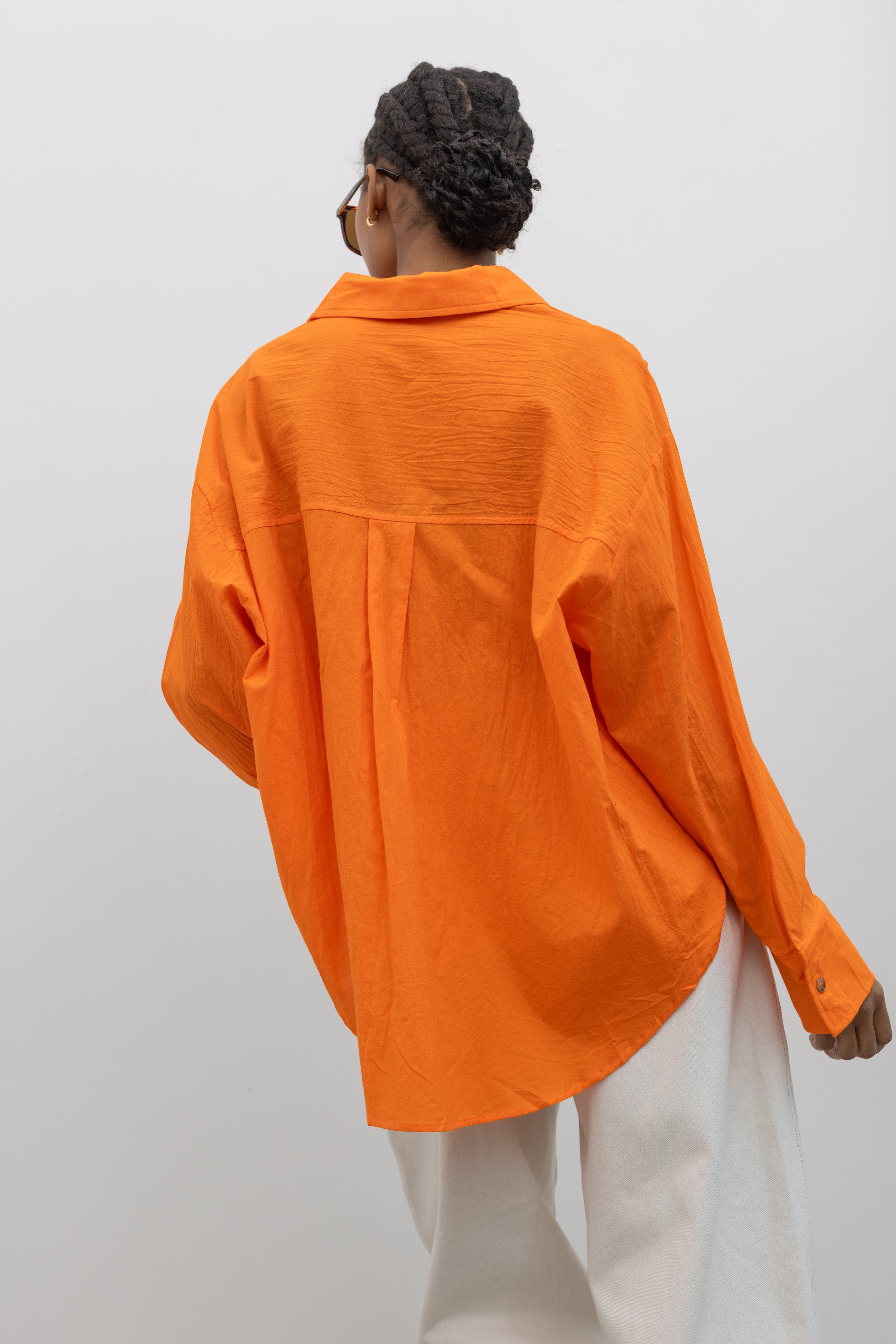 Close-up product back view of a woman wearing Spring/Summer fashion in a Collective tangerine orange cotton oversized button down collared shirt paired with white denim and sunglasses at Alas Eius.