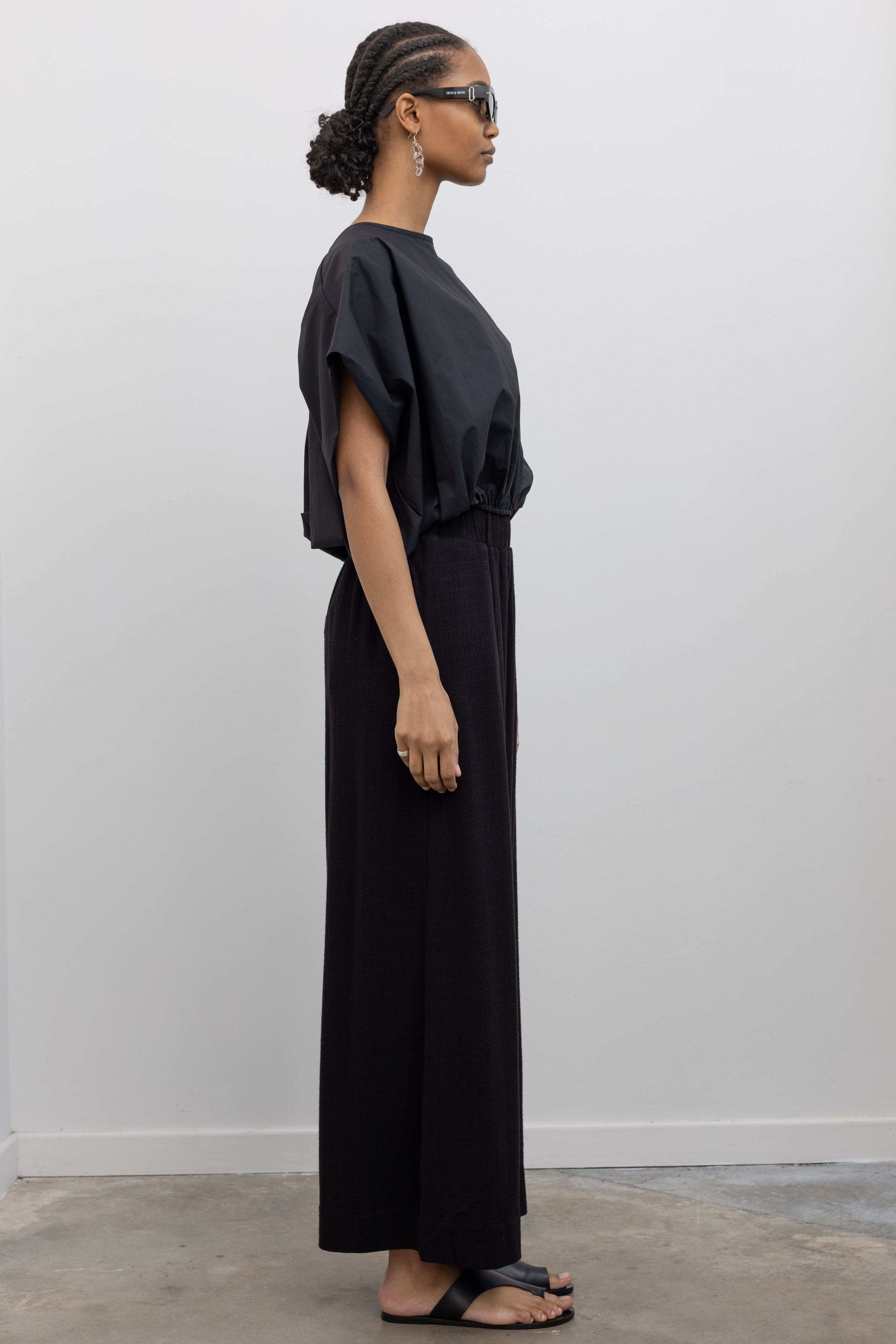 Full length side view of a woman posing wearing elevated spring fashion in the Collective brand black linen wide leg pull on pants with a black balloon hem top and ATP black leather cutout sandals at an Alas Eius store. 