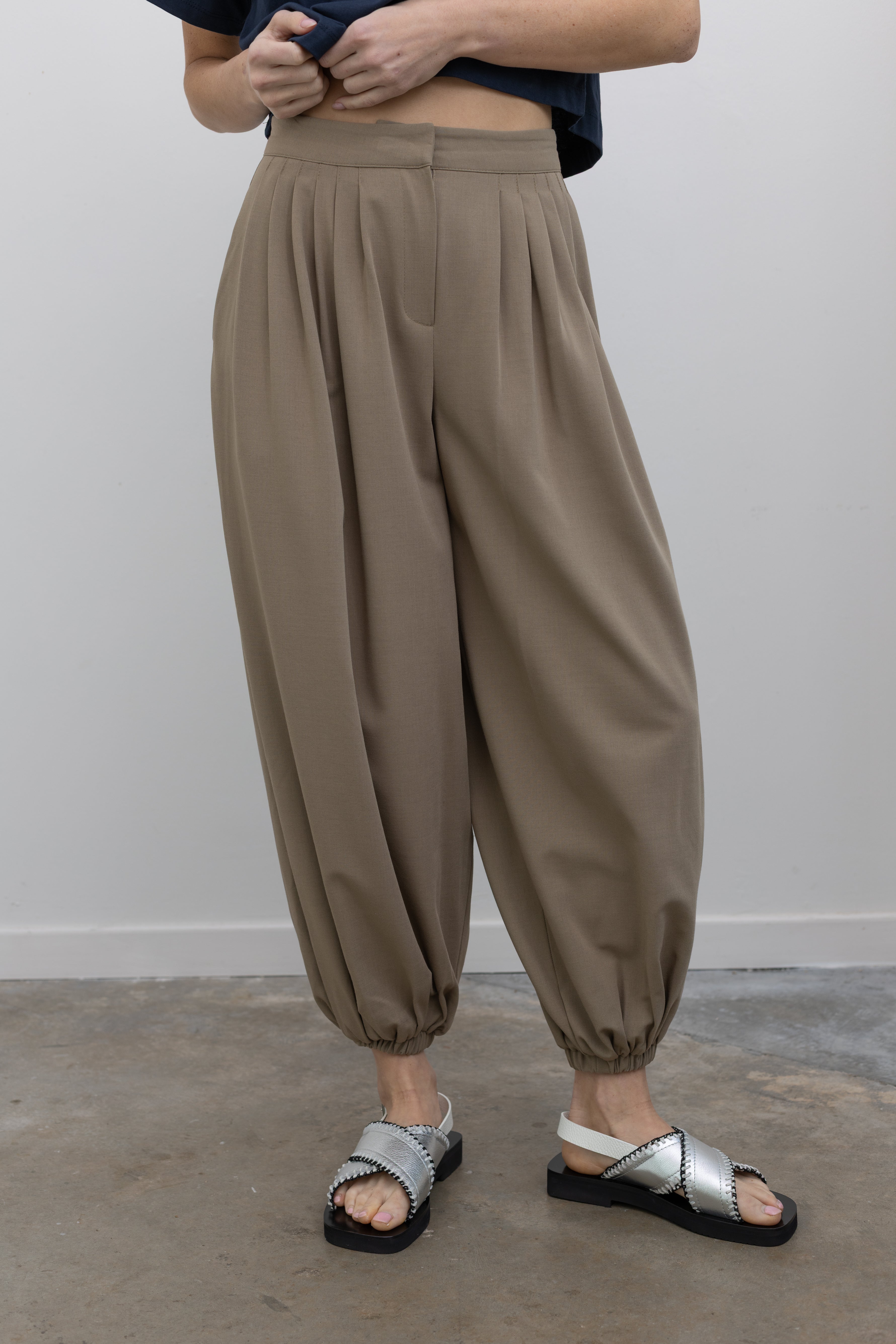 Front view of a woman wearing modern minimalist spring fashion in a Collective brand beige Tores pleated front pant featuring a cinched elastic ankle creating a balloon silhouette paired with an Alas Eius label navy blue t-shirt and metallic silver sandals at an Alas Eius store.