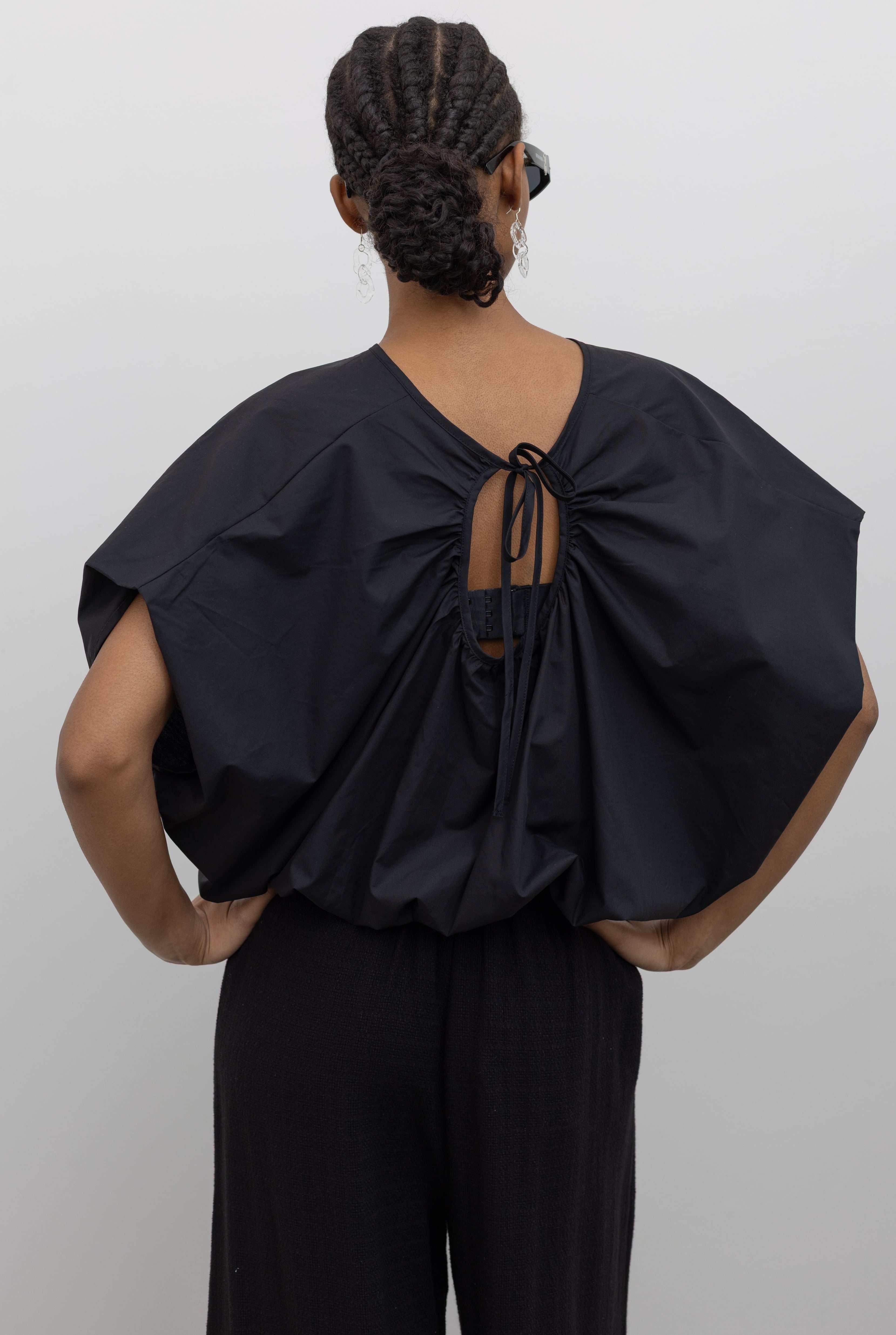 Close-up back view of a woman wearing Spring fashion in a sculptural  Collective Black Cotton billow cinched top featuring draped rounded sleeves, an elasticized waistband for a fitted torso and ribbon tie at the back neck at Alas Eius. 
