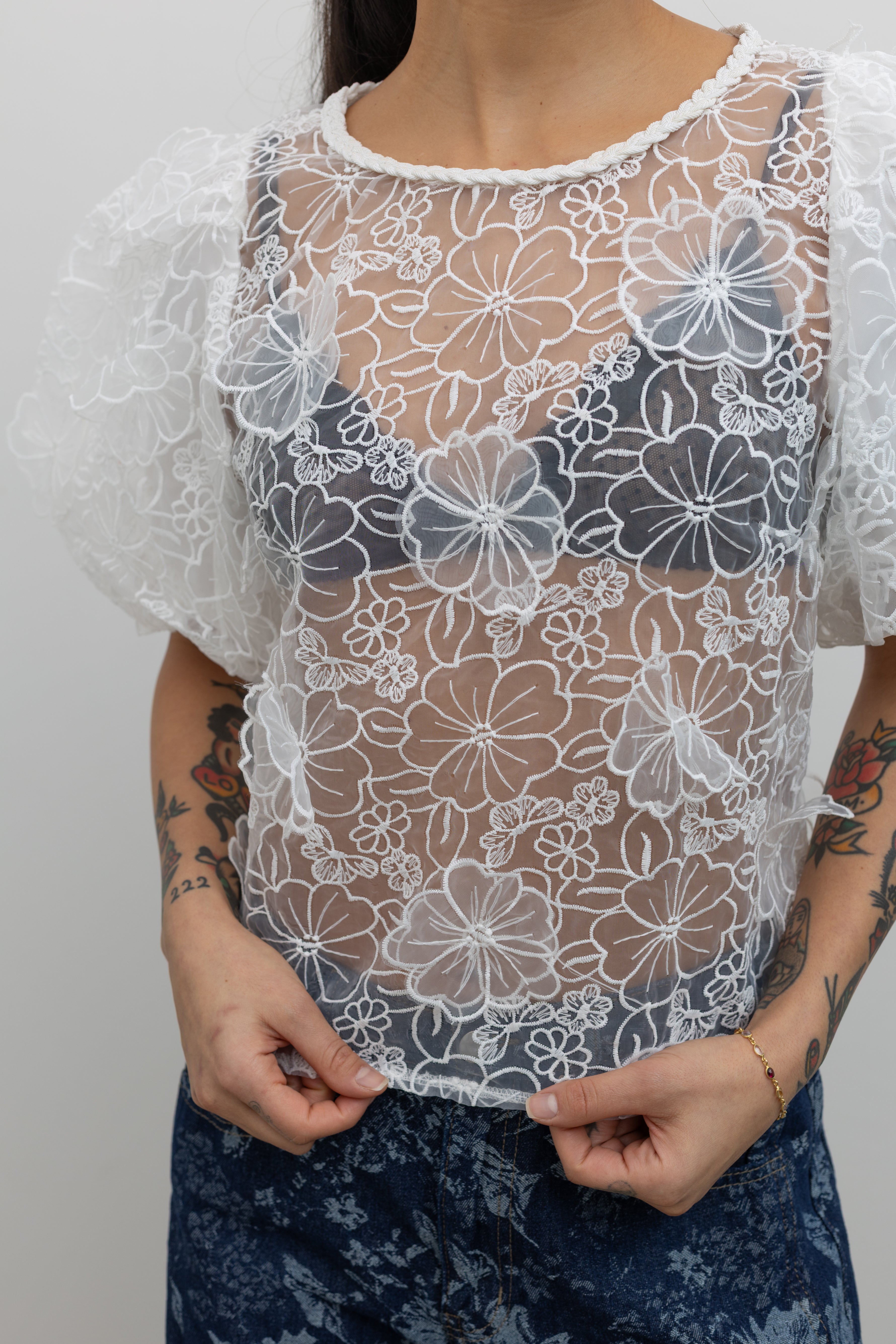 Full length view of a woman posing wearing Spring fashion in a Collective Ivory Floral Embroidered Billow Sleeve Organza Blouse featuring a semi-sheer front paired with floral printed dark wash denim jeans at an Alas Eius store. 