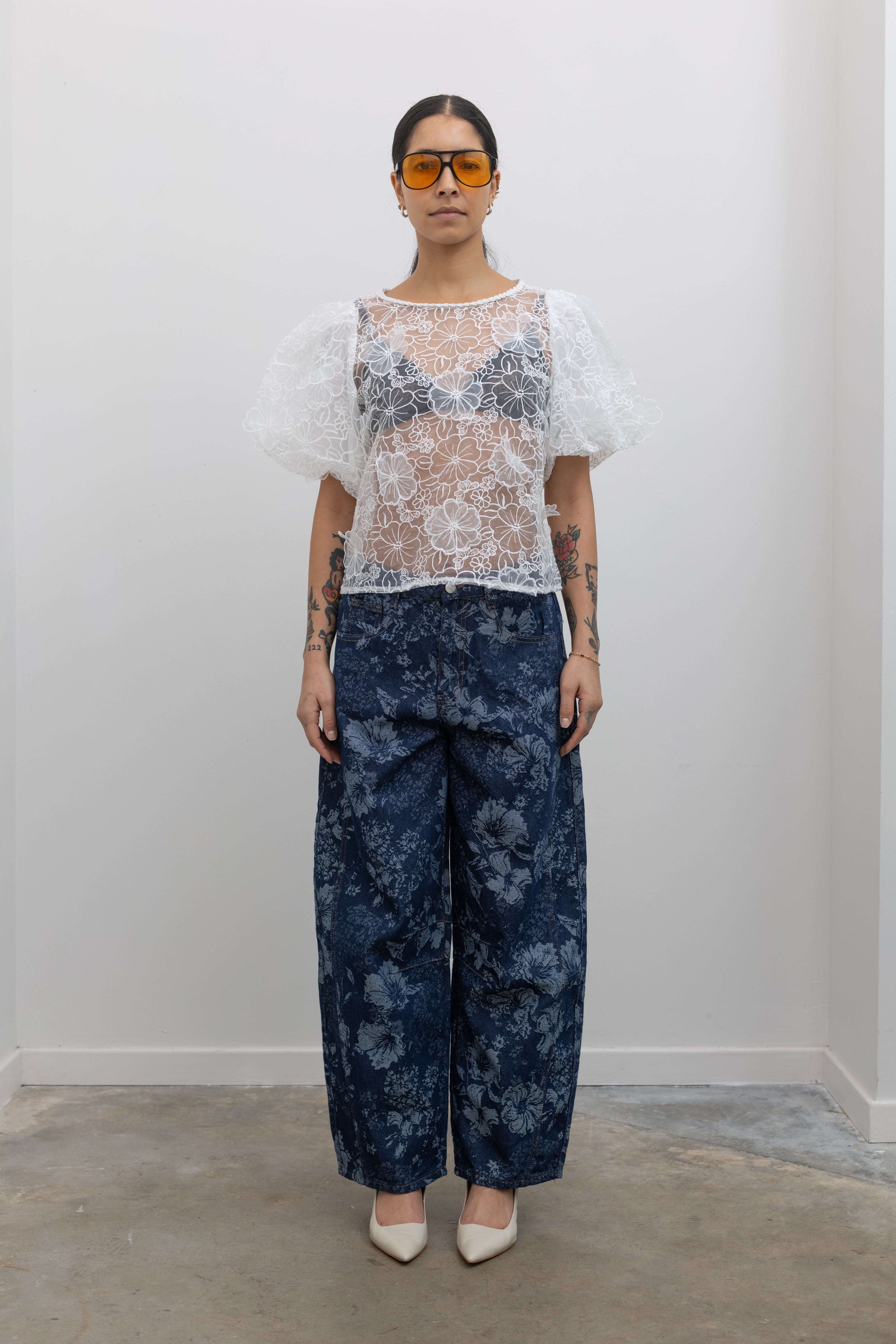 Full length view of a woman posing wearing Spring fashion in a Collective Ivory Floral Embroidered Billow Sleeve Organza Blouse featuring a semi-sheer front paired with floral printed dark wash denim jeans at an Alas Eius store. 
