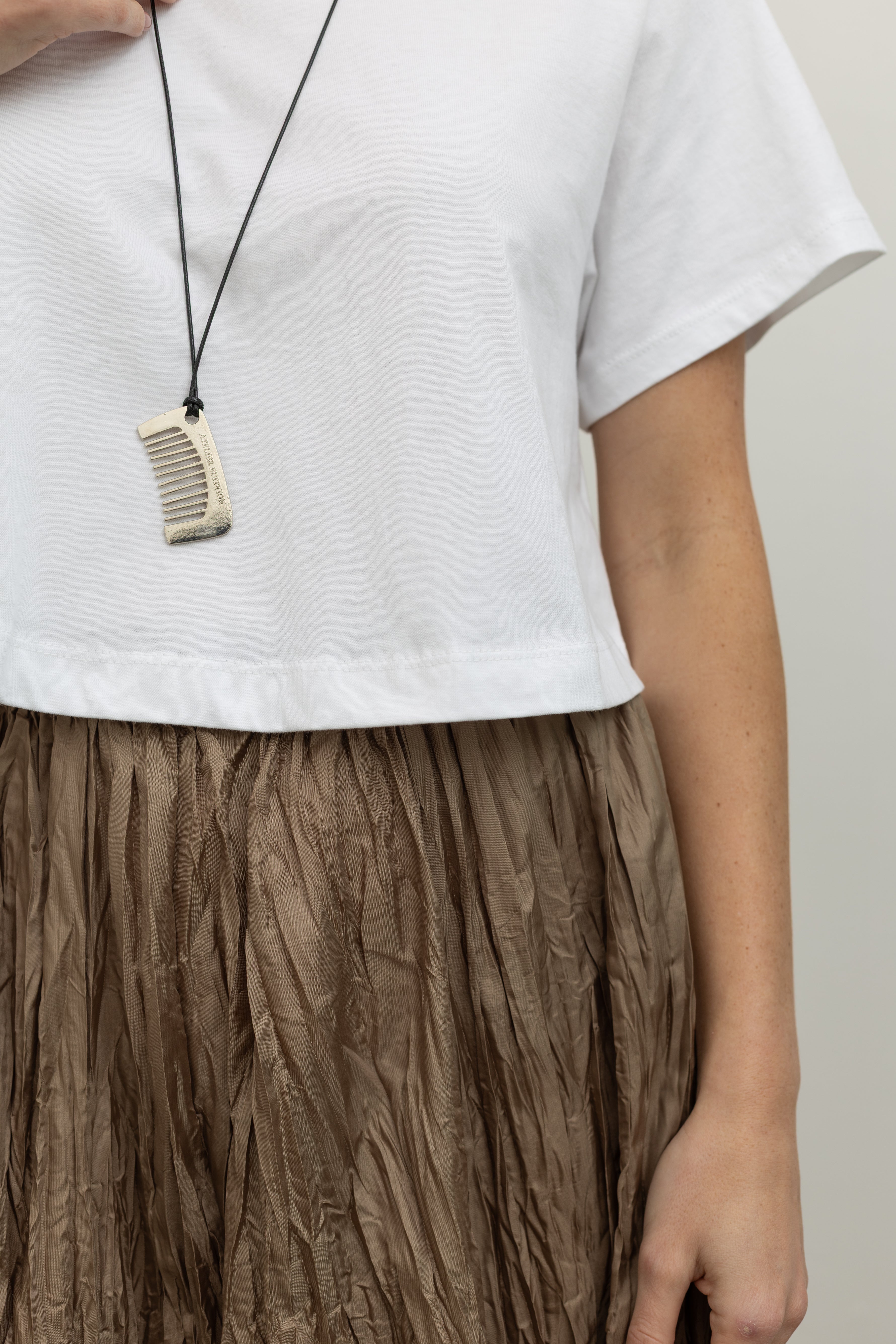 Woman wearing trendy Collective brand stainless steel silver comb pendant on an adjustable leather cord with “Atelier Edition” engraved on one side paired with an Alas Eius label white cotton cropped t-shirt, sunglasses, and crinkled maxi skirt at an Alas Eius store.