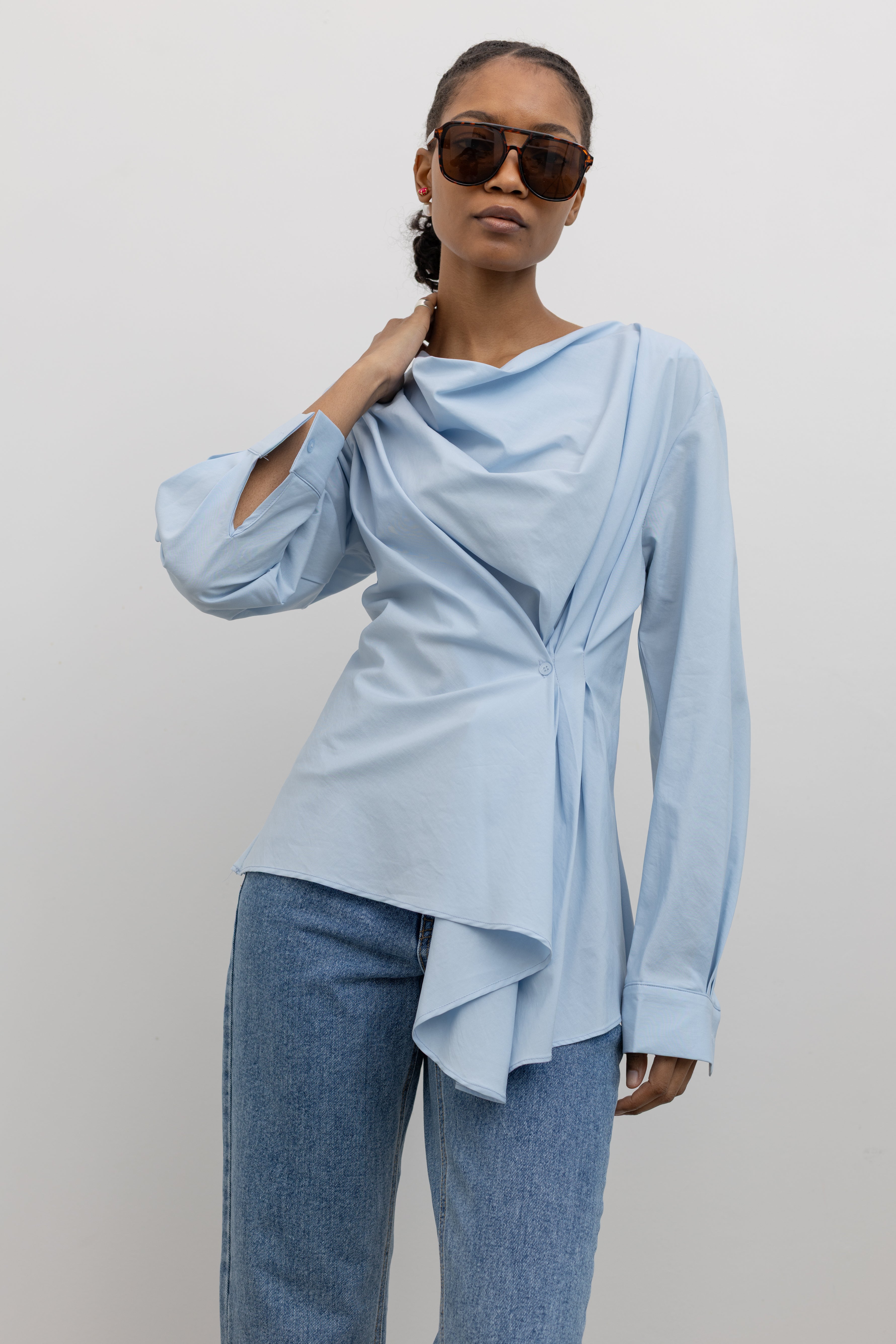 Woman wearing chic and architectural spring fashion in a Collective label sky blue cotton long sleeve top featuring a fitted natural waistline with a side draped motif creating an asymmetrical hem and cowl neckline paired with light wash denim jeans at Alas Eius. 