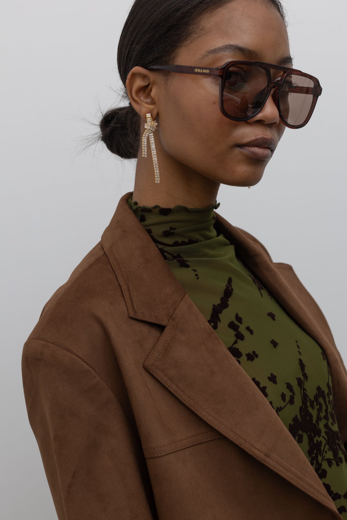 Woman posing wearing sophisticated contemporary women's fashion in a Collective label olive green mesh high neck lettuce hem top featuring a raised floral printed throughout paired with a brown suede jacket at an Alas Eius store. 