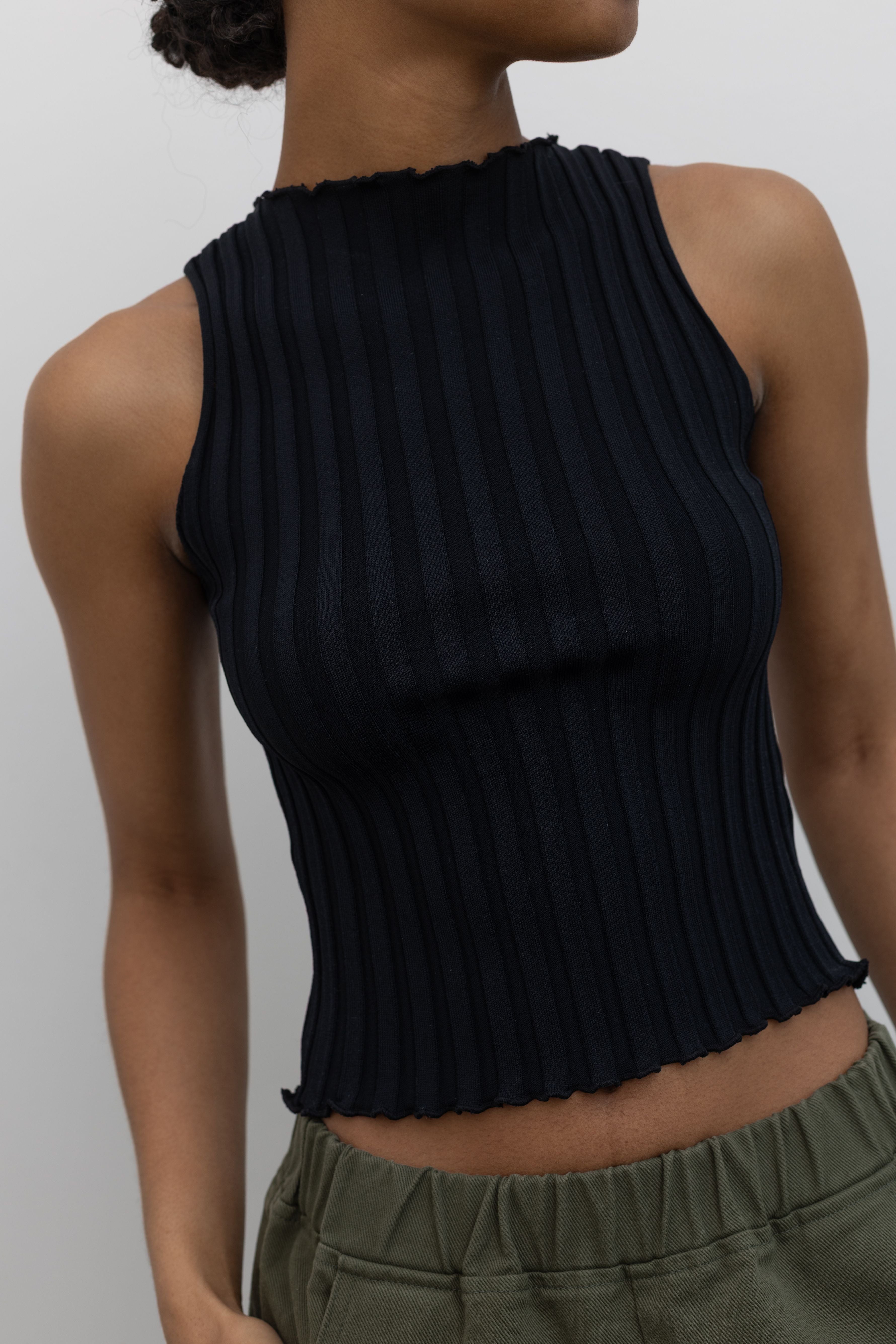 Close-up front detailed view of a woman wearing spring capsule wardrobe essential in a Collective brand black fitted ribbed knit lettuce hem sleeveless top with slight mock neck paired with Le Bon Shoppe olive green cotton canvas arc shaped pant at Alas Eius. 