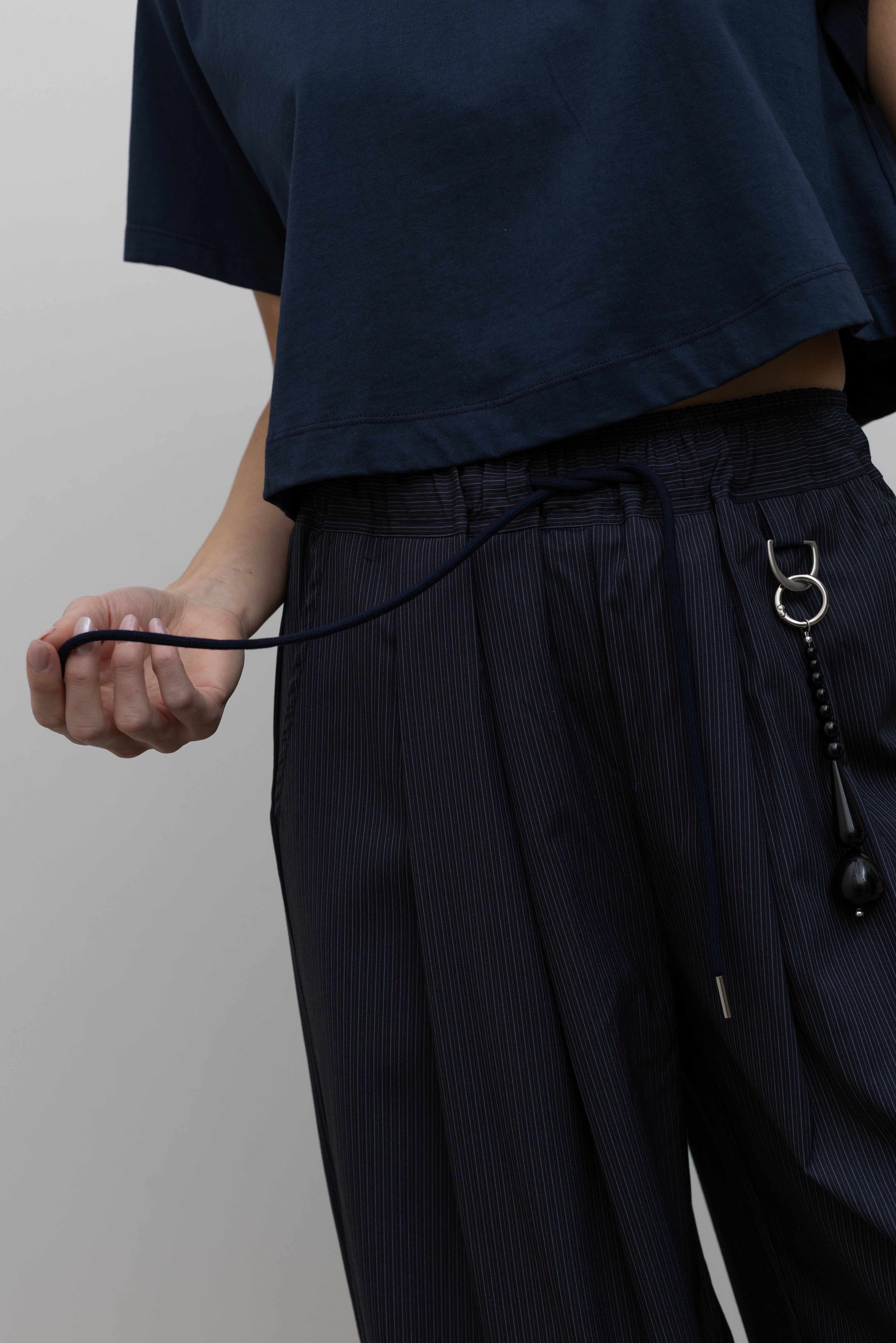 Detailed product view of Woman standing wearing Spring fashion in a pair of Collective Navy Pinstripe Wide Leg Drawstring Pants and navy blue tee with black beaded accessory at belt loop at Alas Eius