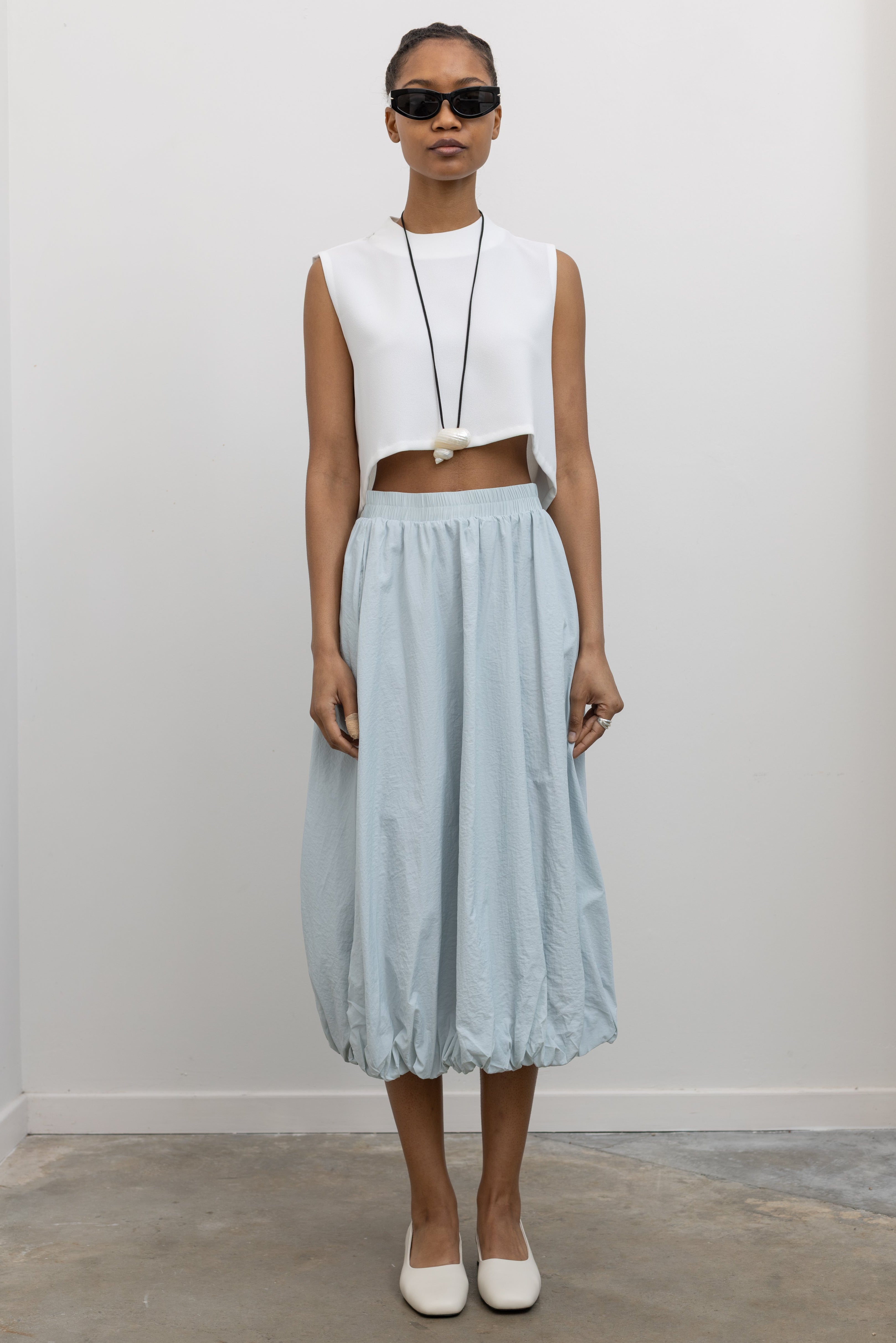 Woman standing wearing Spring fashion in a Collective Light Blue Elastic Waist Bubble Hem Midi Skirt with a white sleeveless top and white shoes at Alas Eius