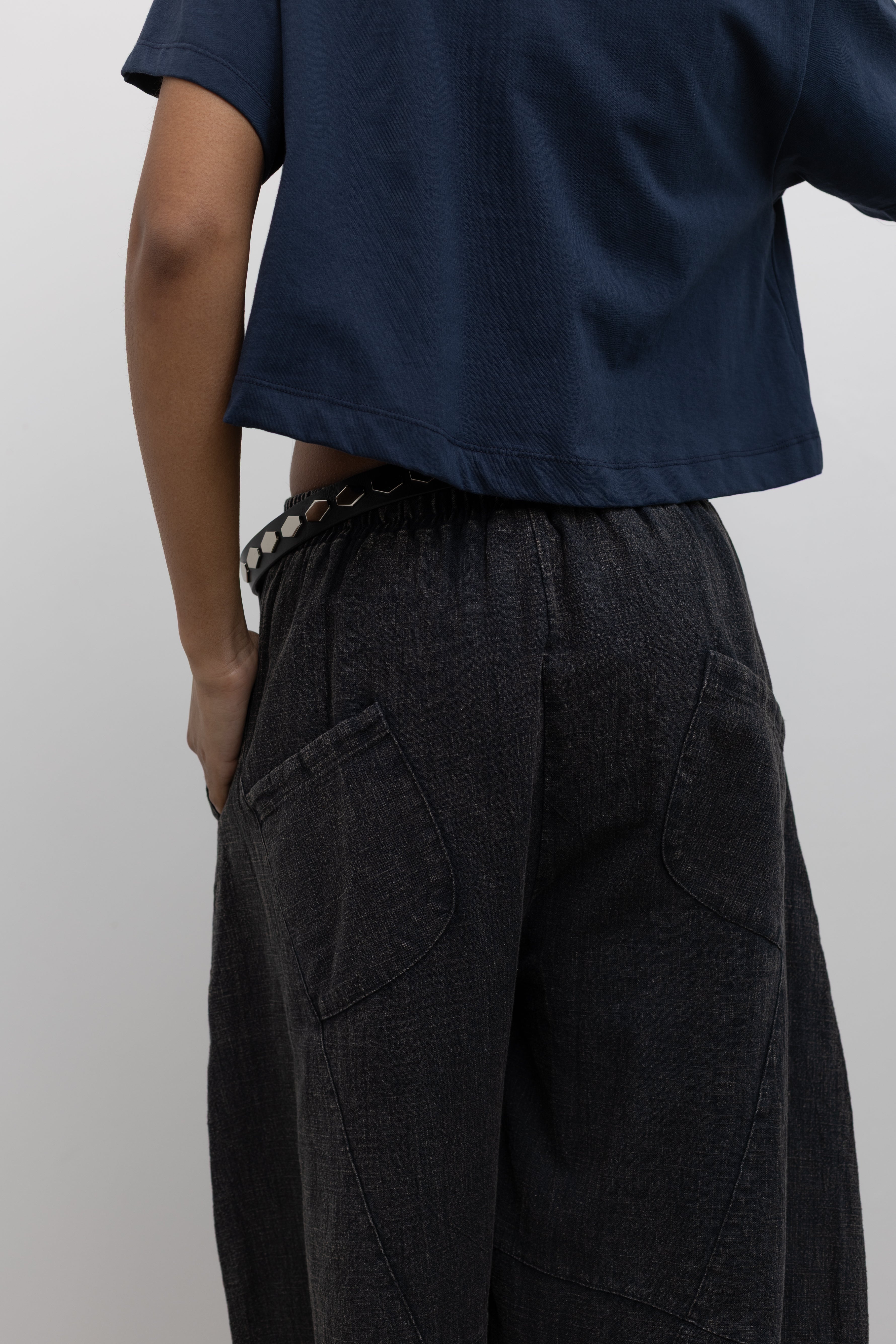 Close-up back view of a woman wearing spring contemporary fashion in a Alas Eius label navy blue cropped t-shirt paired with Collective Spring Black Barrel Cotton Blend Pants at Alas Eius. 