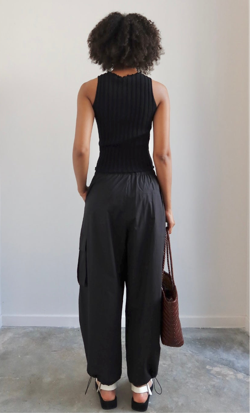 Close-up back view of a woman wearing spring capsule wardrobe essential in a Collective brand black fitted ribbed knit lettuce hem sleeveless top with slight mock neck paired with Alas Eius label black cargo pant and Flattered white leather fisherman platform buckle sandals at Alas Eius. 