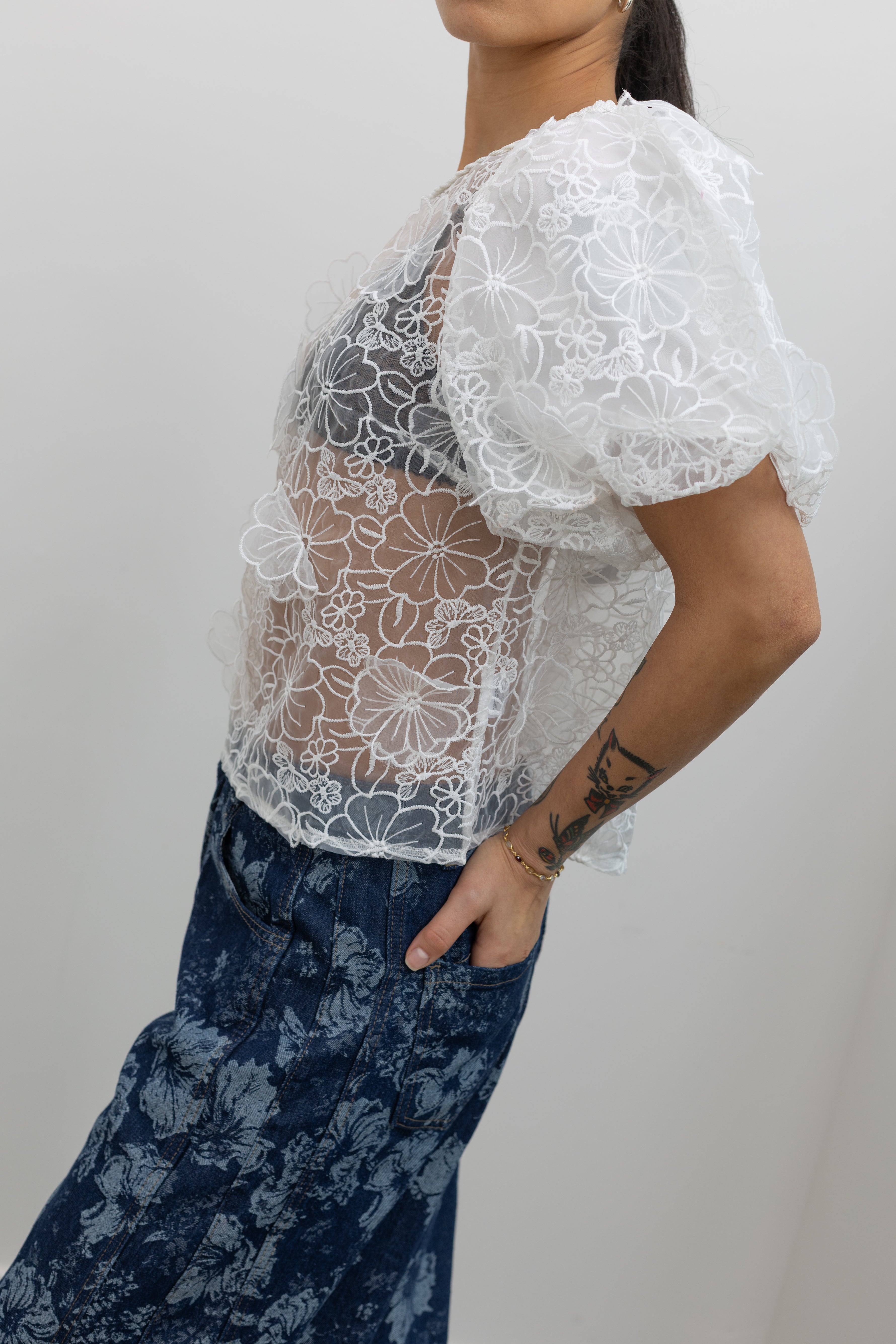 Side view of a woman posing wearing Spring fashion in a Collective Ivory Floral Embroidered Billow Sleeve Organza Blouse featuring a semi-sheer front paired with floral printed dark wash denim jeans at an Alas Eius store. 
