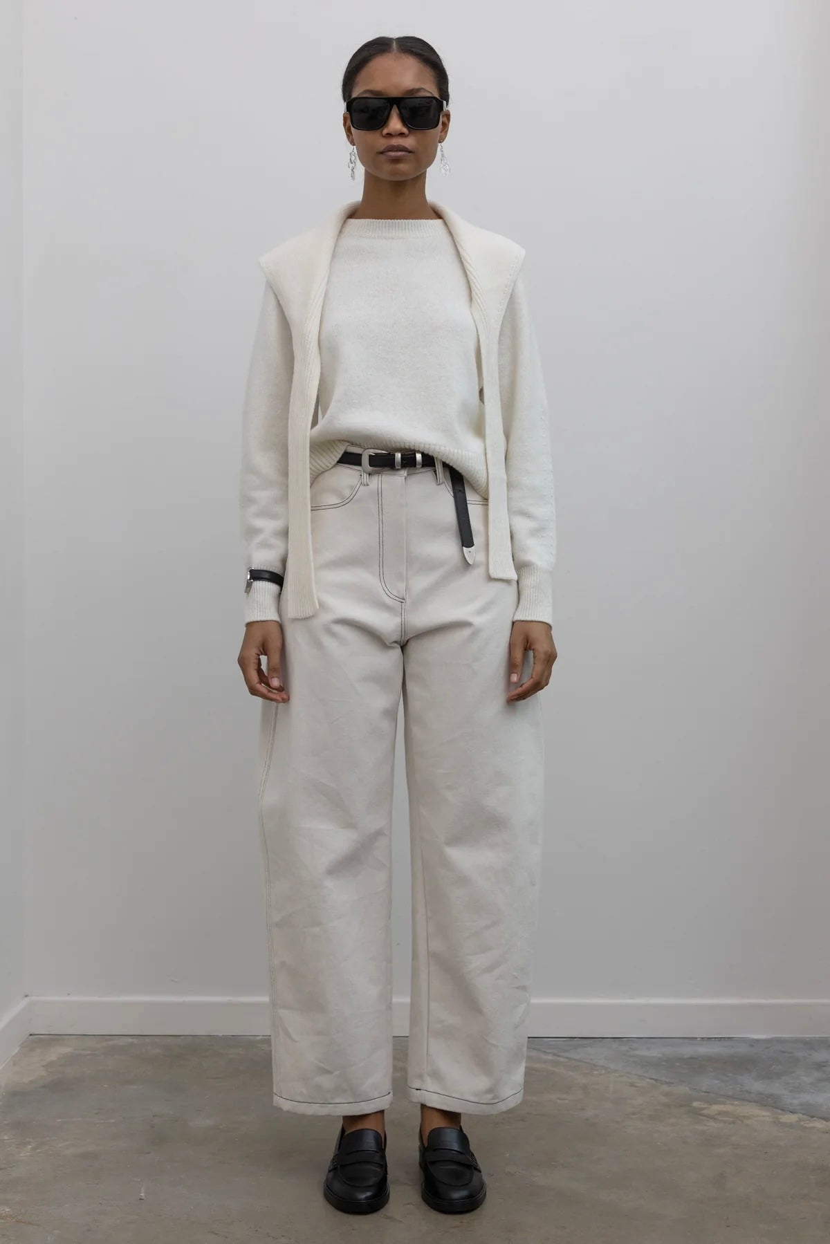 Full length front view of a woman standing wearing an everyday capsule wardrobe outfit in a white long sleeve top with Collective Spring Cream Curved leg pants featuring Contrasting stitching paired with a black leather belt with silver hardware and black leather loafers at Alas Eius. 