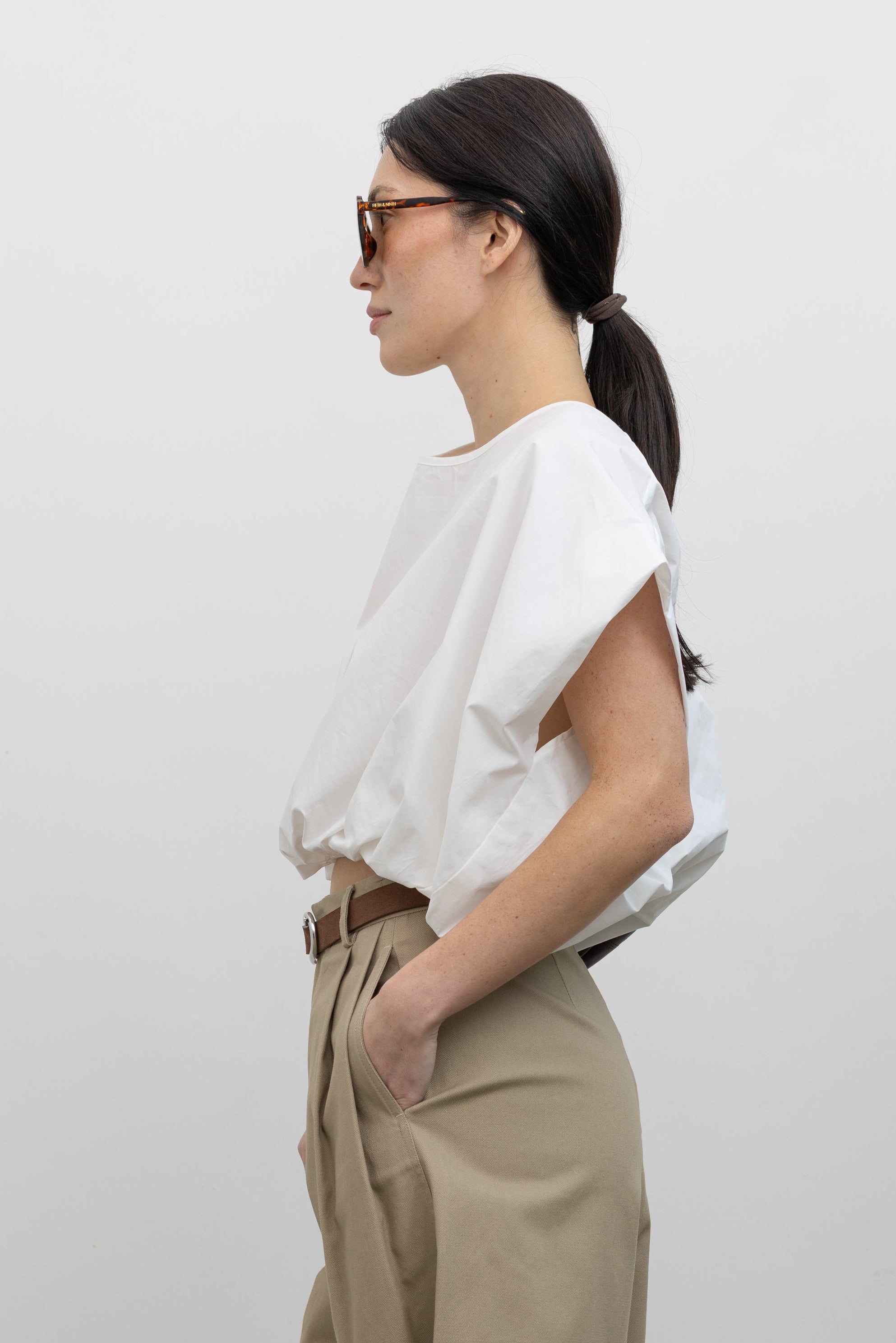 Close-up side view of a woman wearing Spring fashion in a sculptural  Collective White Cotton billow cinched top featuring draped rounded sleeves, an elasticized waistband for a fitted torso and ribbon tie at the back neck paired with timeless khaki pleated high waisted trousers and a brown leather belt at Alas Eius. 