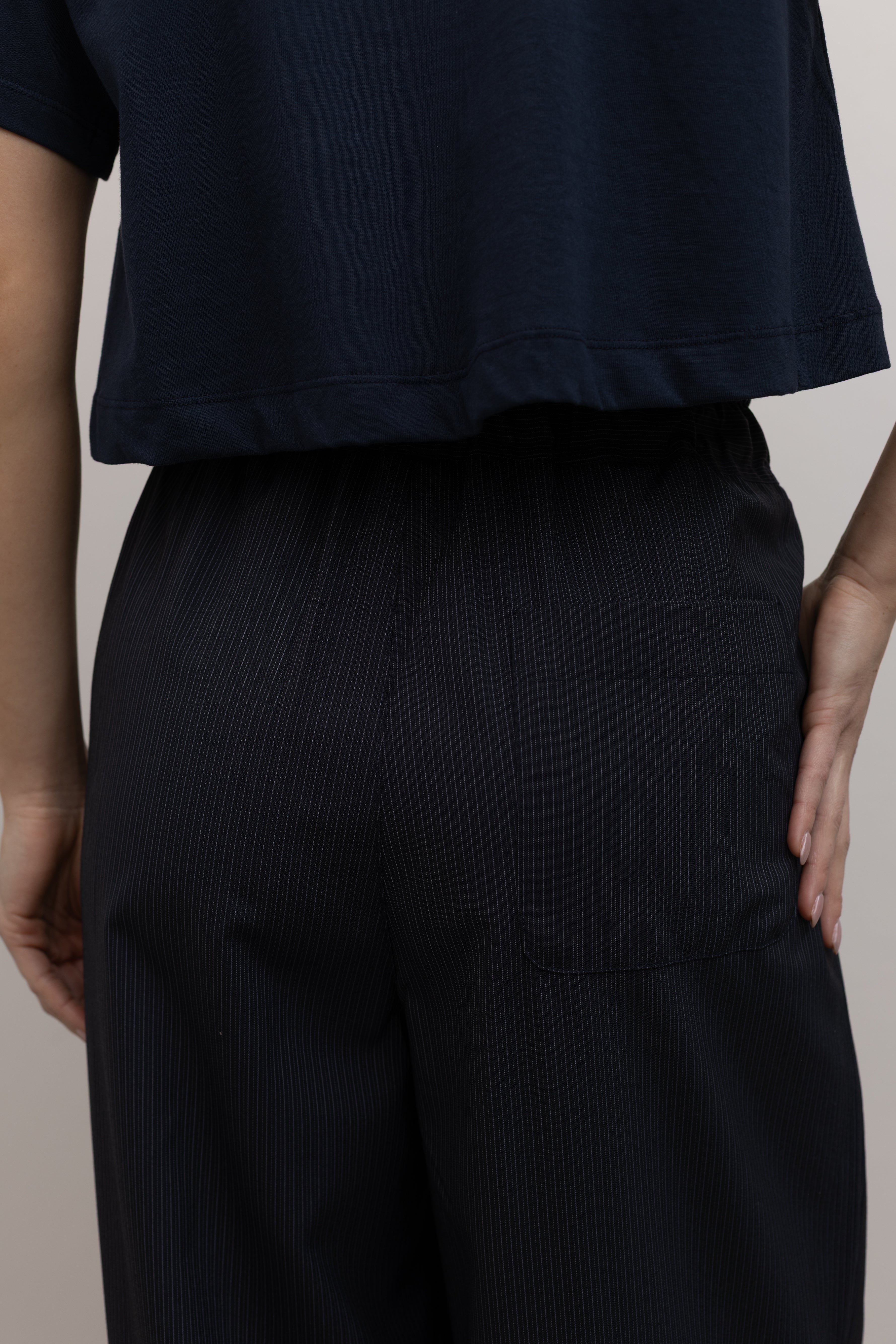 Detailed product back view of Woman standing wearing Spring fashion in a pair of Collective Navy Pinstripe Wide Leg Drawstring Pants and navy blue tee with black beaded accessory at belt loop at Alas Eius