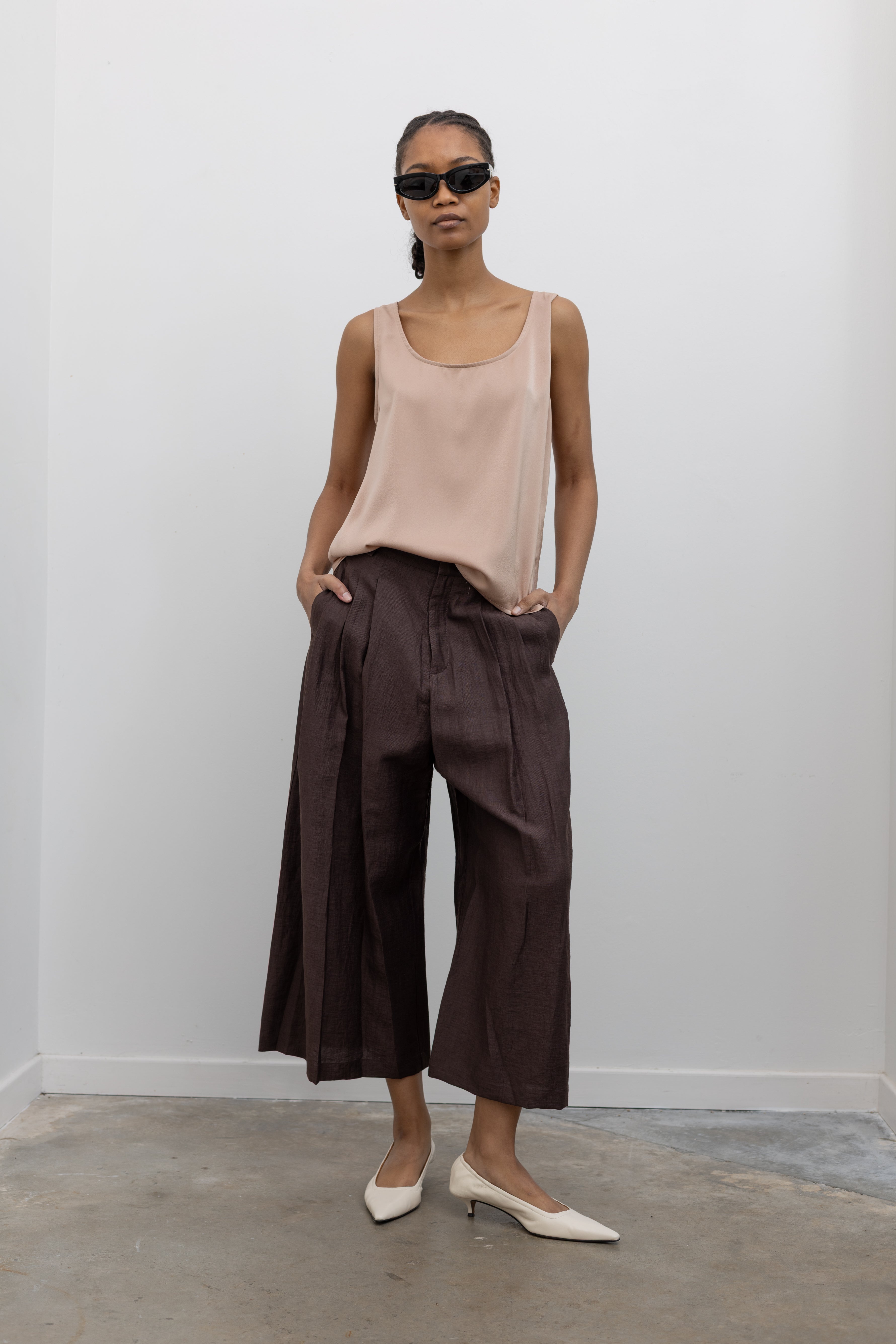 Front view of a woman posing with hands in her pockets wearing Spring fashion in a Collective brand solid beige satin tank top with a relaxed flowy fit paired with pleated trouser at Alas Eius.
