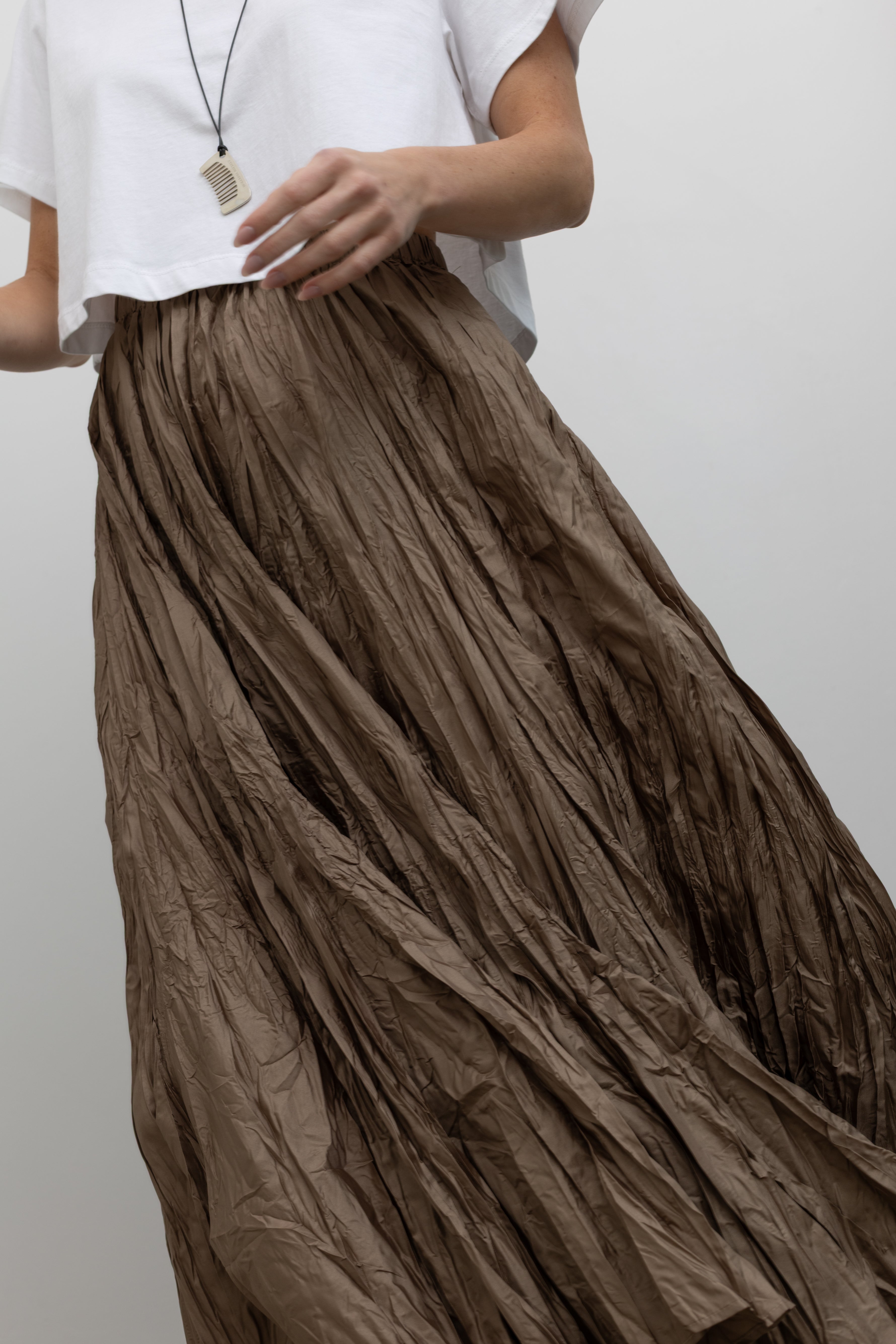 Close-up front view of a woman wearing contemporary spring fashion in an Alas Eius label cropped white t-shirt paired with a Collective Khaki Crinkle Textured Maxi Skirt at Alas Eius. 
