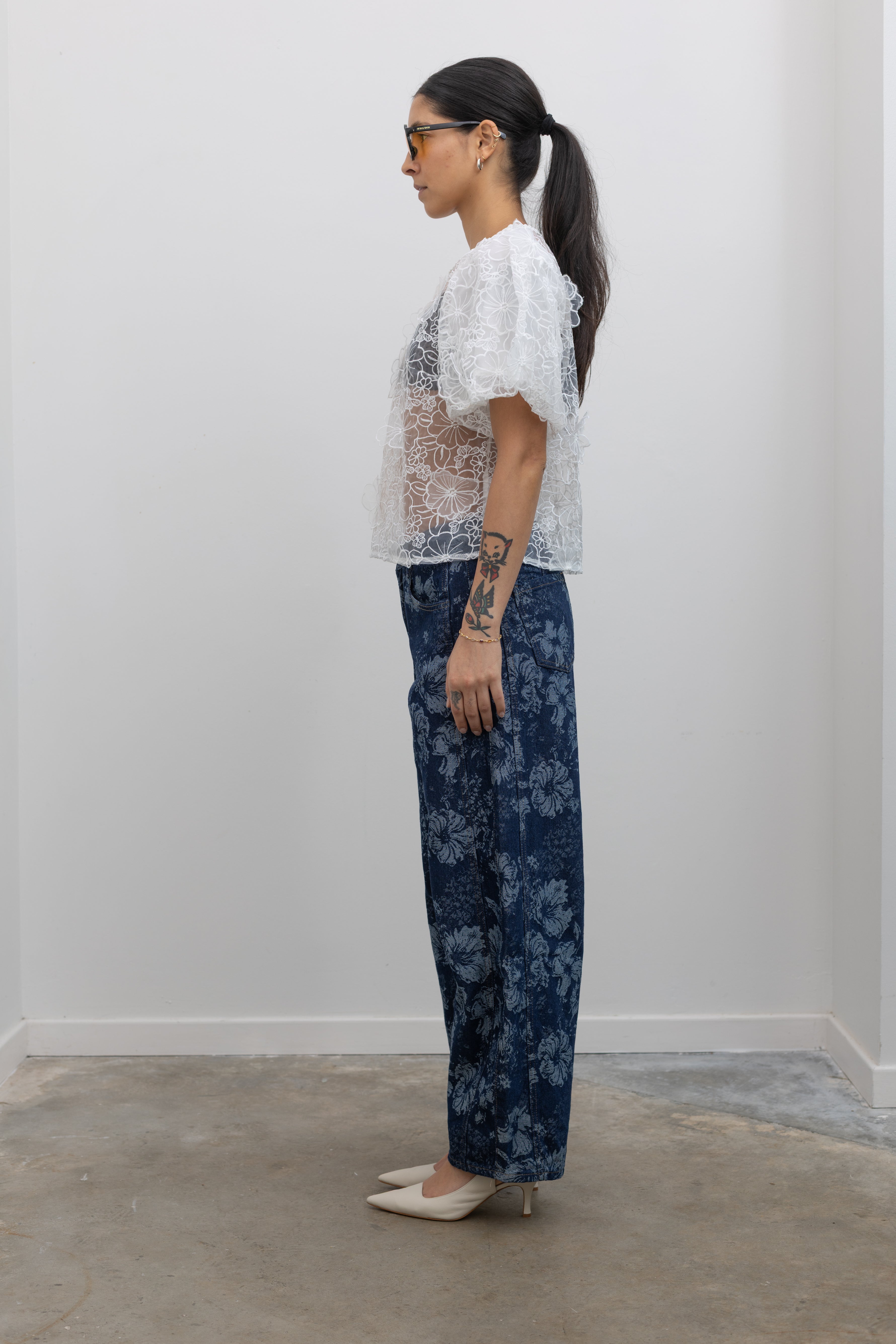 Full length side view of a woman posing wearing Spring fashion in a Collective Ivory Floral Embroidered Billow Sleeve Organza Blouse featuring a semi-sheer front paired with floral printed dark wash denim jeans at an Alas Eius store. 