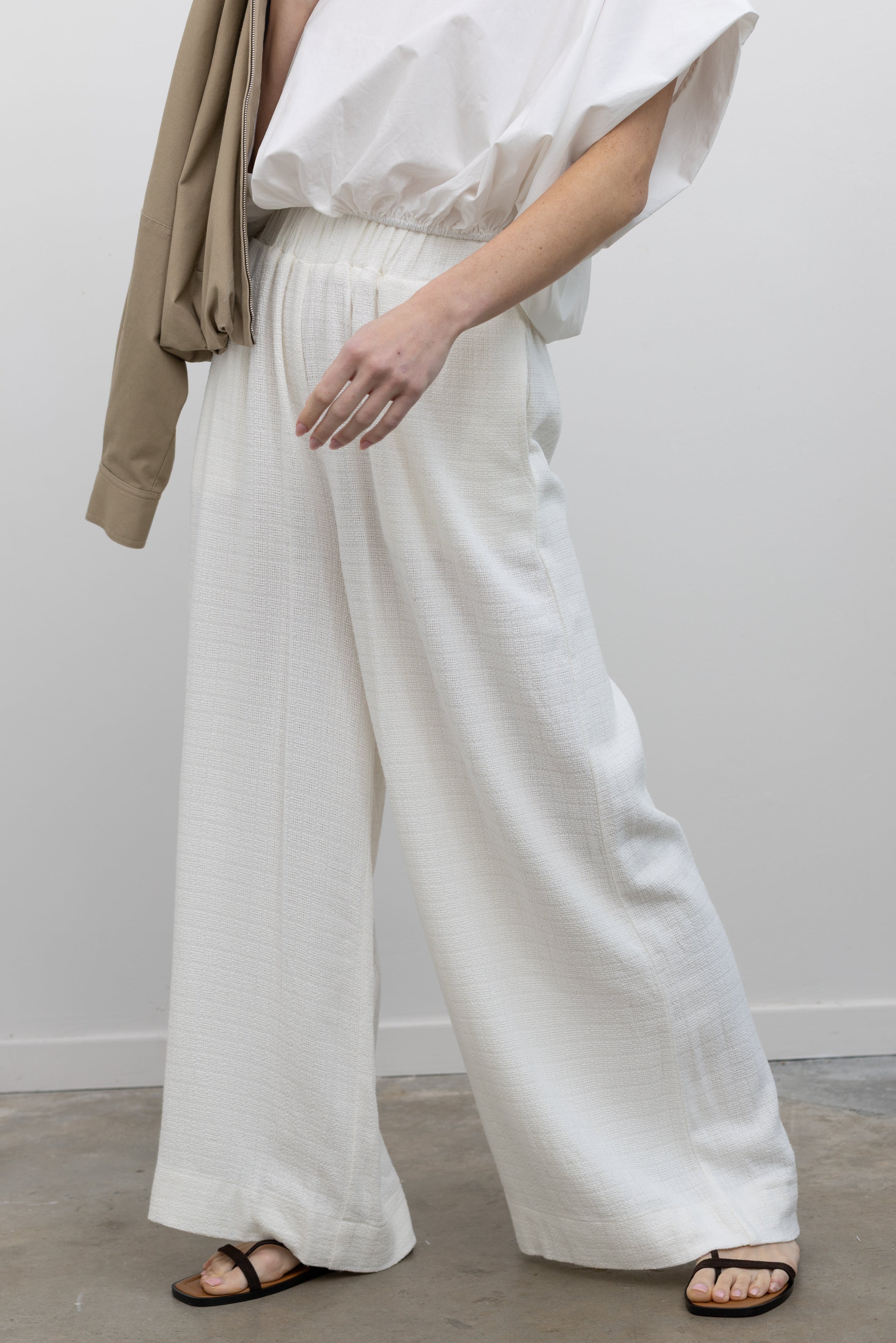 Close-up view of woman in motion wearing chic spring fashion in Collective Ivory Linen Wide Leg Pants featuring an elastic waistband and side seam pockets paired with an oversized short sleeve top and strappy leather sandals at Alas Eius.