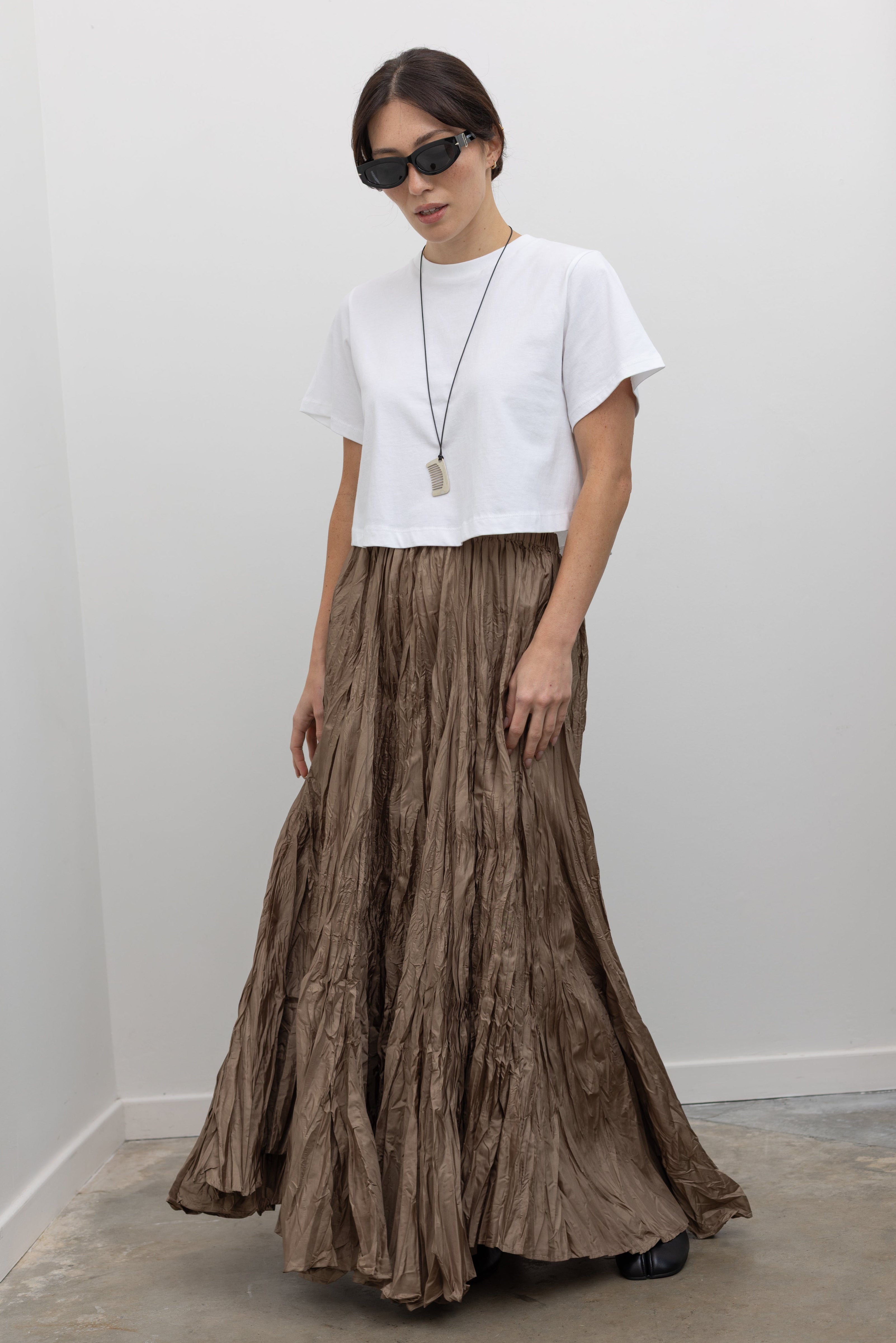 Full length front view of a woman standing wearing elevated spring fashion in an Alas Eius label cropped white t-shirt paired with a Collective Khaki Crinkle Textured Maxi Skirt at Alas Eius. 