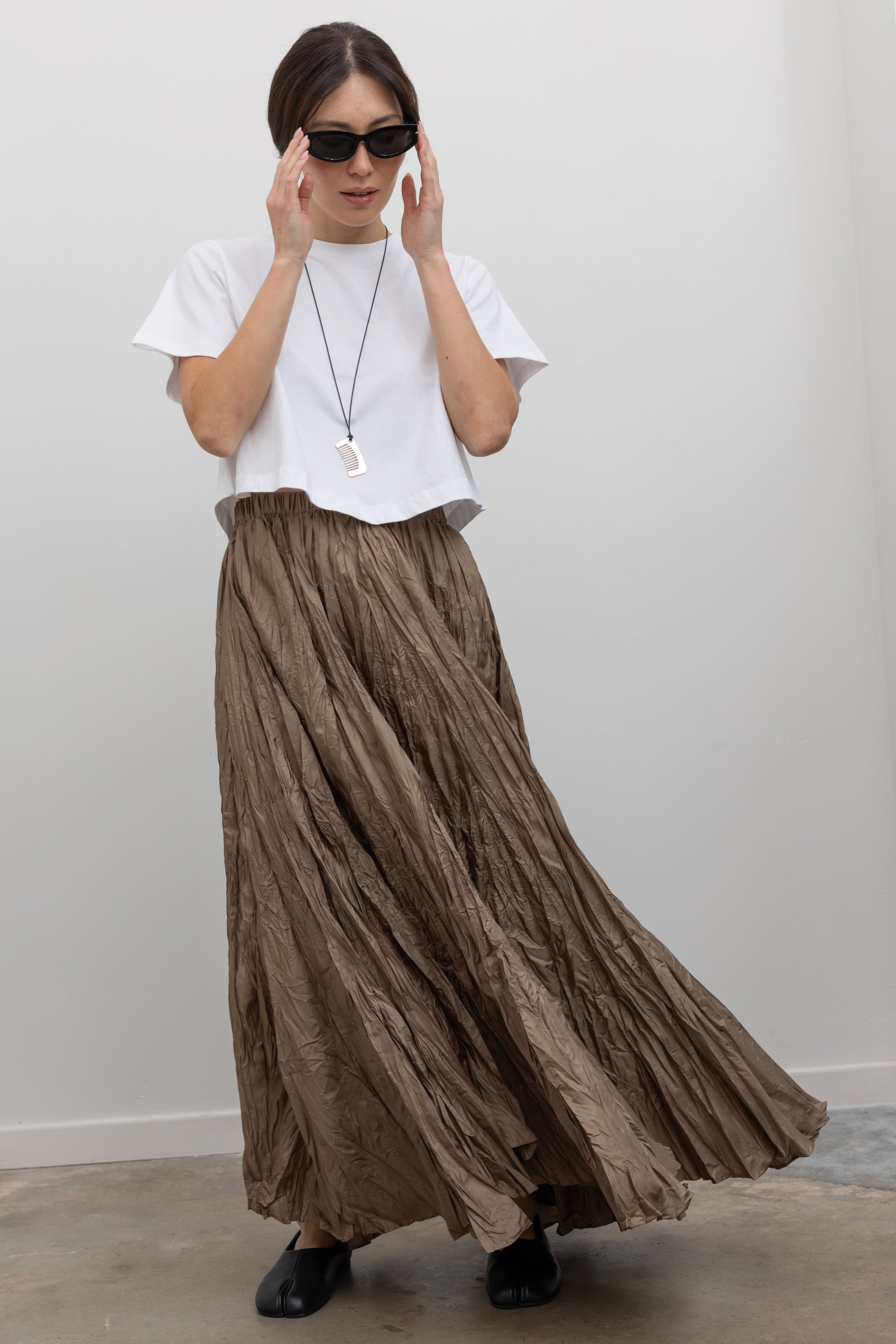 Full length front view of a woman in motion wearing elevated spring fashion in an Alas Eius label cropped white t-shirt paired with a Collective Khaki Crinkle Textured Maxi Skirt at Alas Eius. 