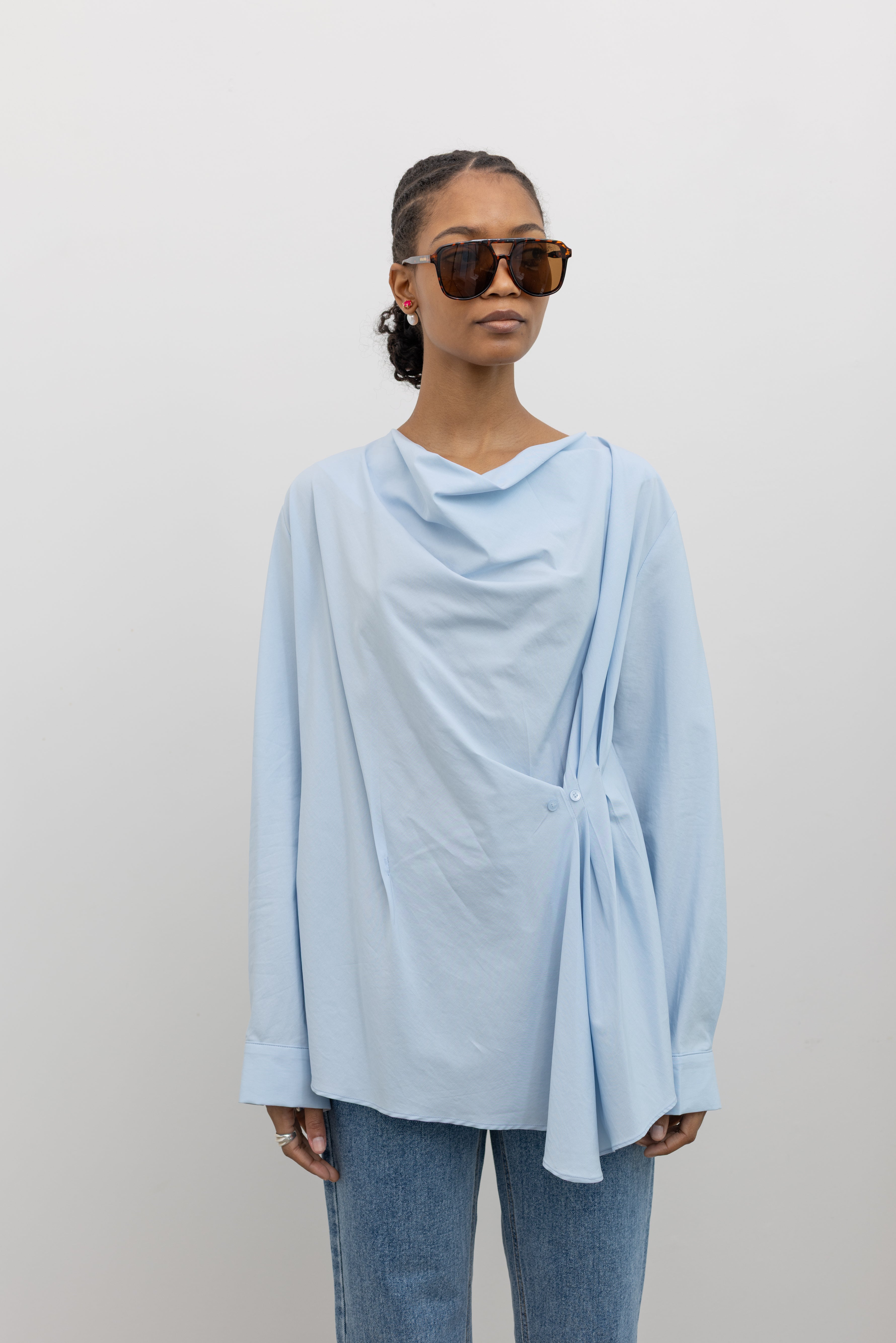 Woman wearing chic and architectural spring fashion in a Collective label sky blue cotton long sleeve top featuring a fitted natural waistline with a side draped motif creating an asymmetrical hem and cowl neckline paired with light wash denim jeans at Alas Eius. 