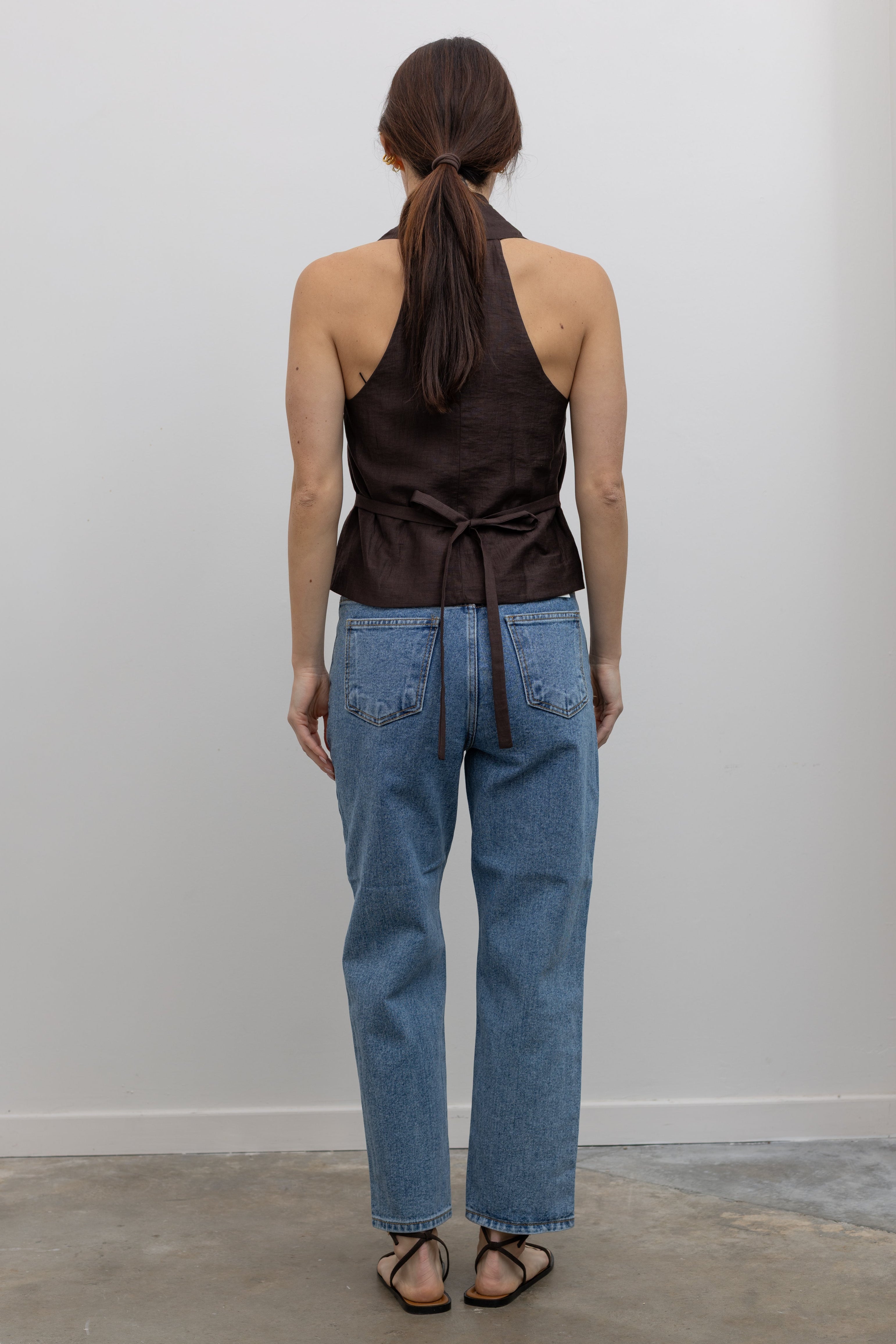 Back view of Woman standing wearing Spring fashion in a brown linen pleated vest and a pair of Collective Stone Wash Carrot High Rise Jeans with strappy sandals and sunglasses at Alas Eius