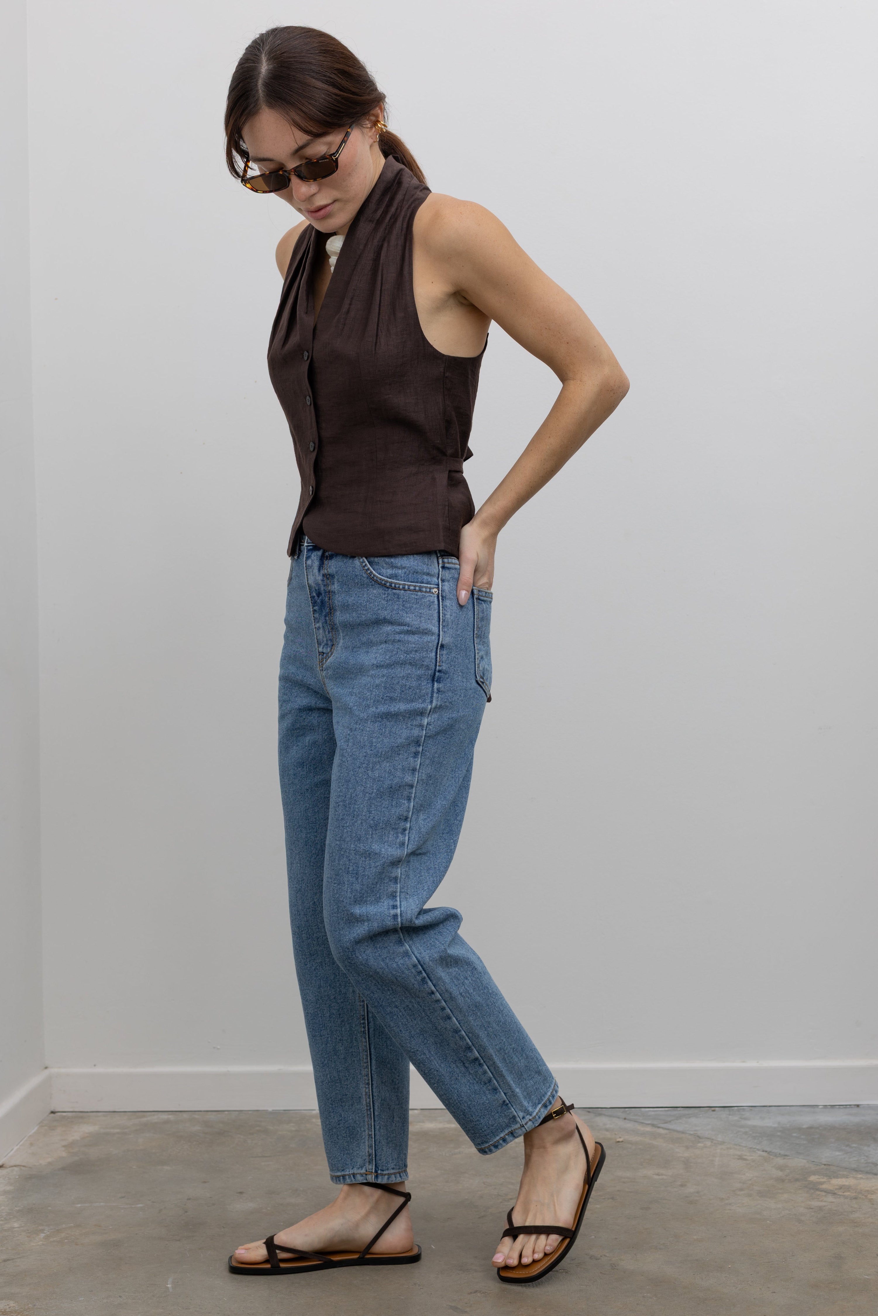 Side view of Woman standing wearing Spring fashion in a brown linen pleated vest and a pair of Collective Stone Wash Carrot High Rise Jeans with strappy sandals and sunglasses at Alas Eius