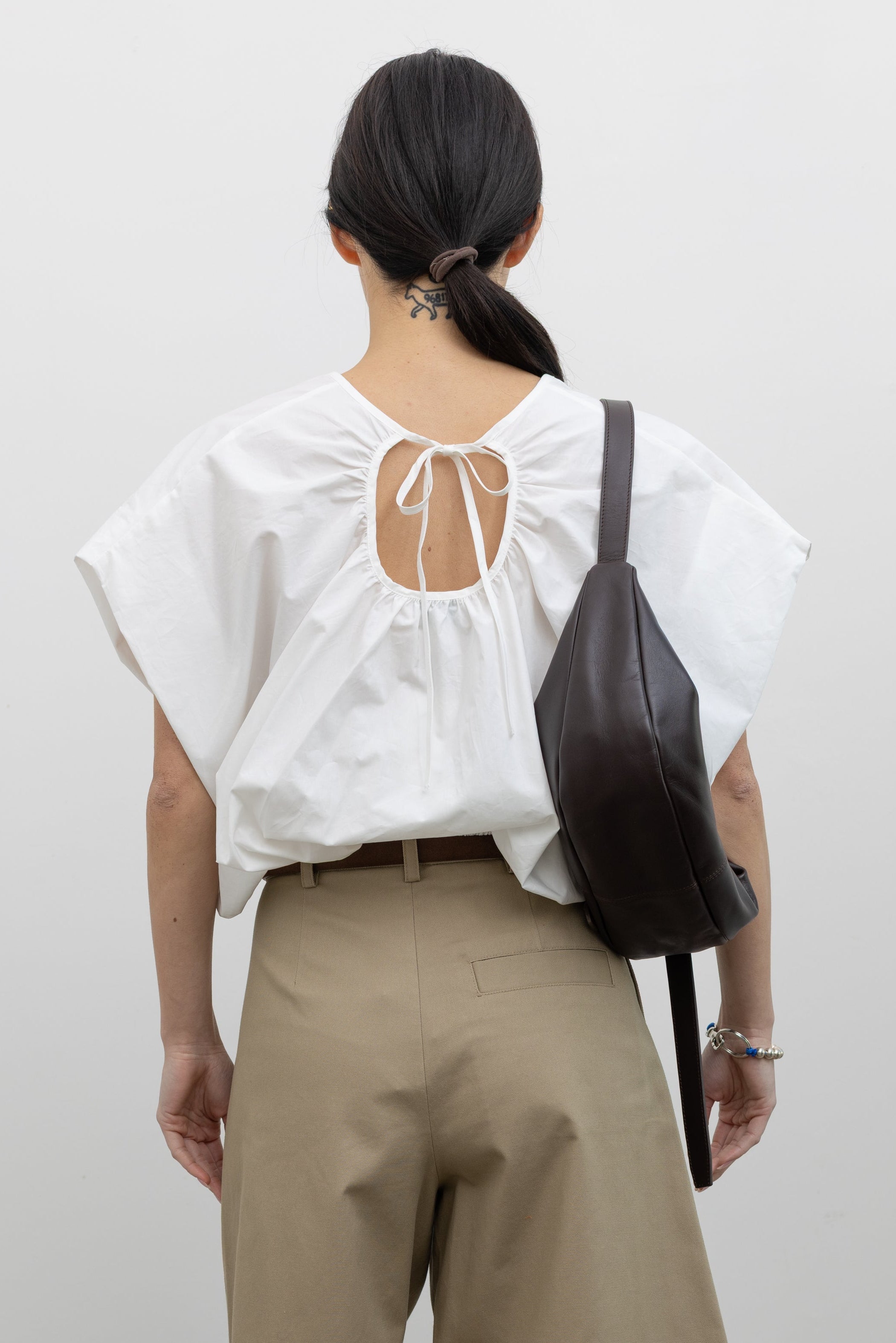 Close-up back view of a woman wearing Spring fashion in a sculptural  Collective White Cotton billow cinched top featuring draped rounded sleeves, an elasticized waistband for a fitted torso and ribbon tie at the back neck paired with timeless khaki pleated high waisted trousers and a brown leather belt at Alas Eius. 