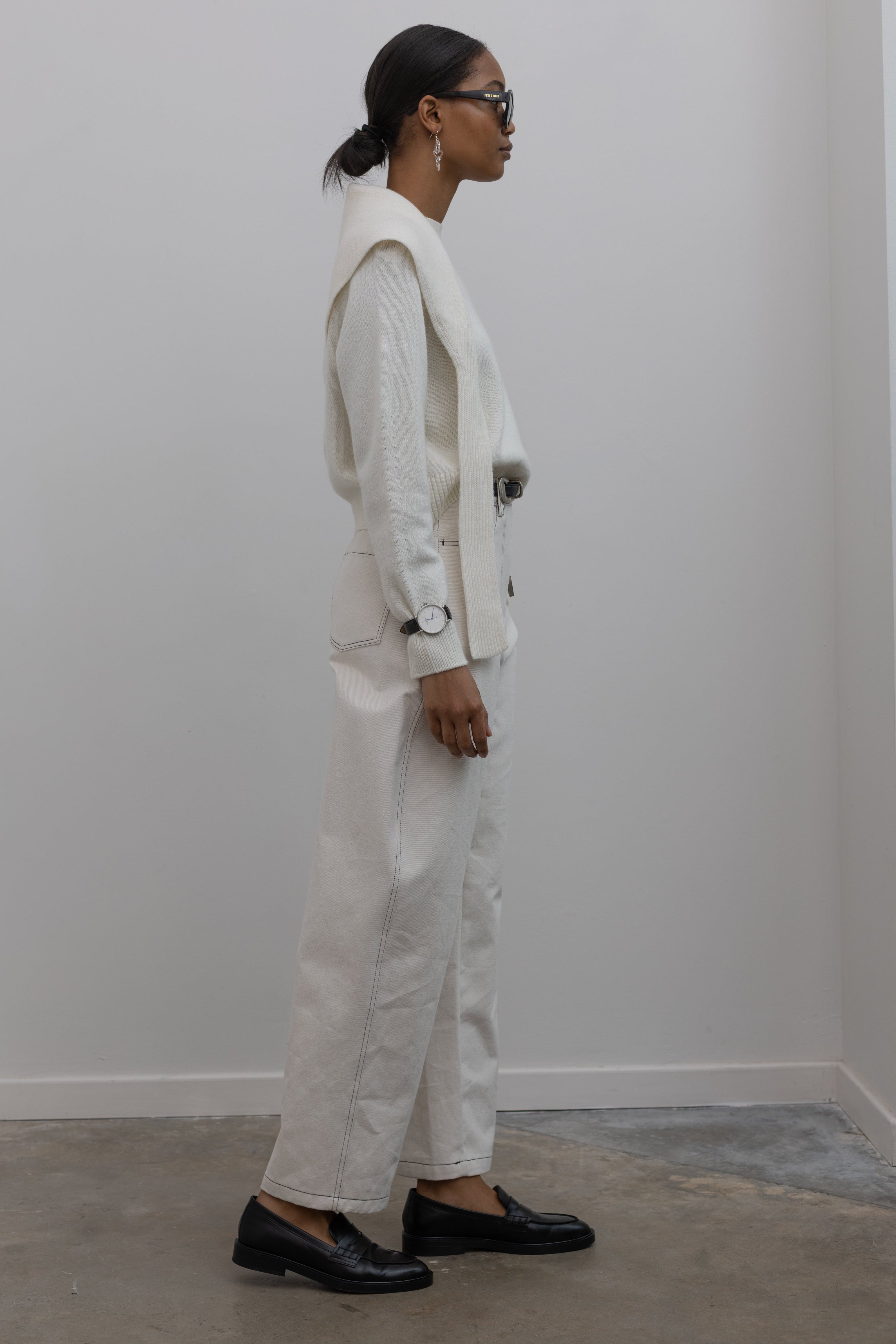 Full length side view of a woman standing wearing an everyday capsule wardrobe outfit in a white long sleeve top with Collective Spring Cream Curved leg pants featuring Contrasting stitching paired with a black leather belt with silver hardware and black leather loafers at Alas Eius. 