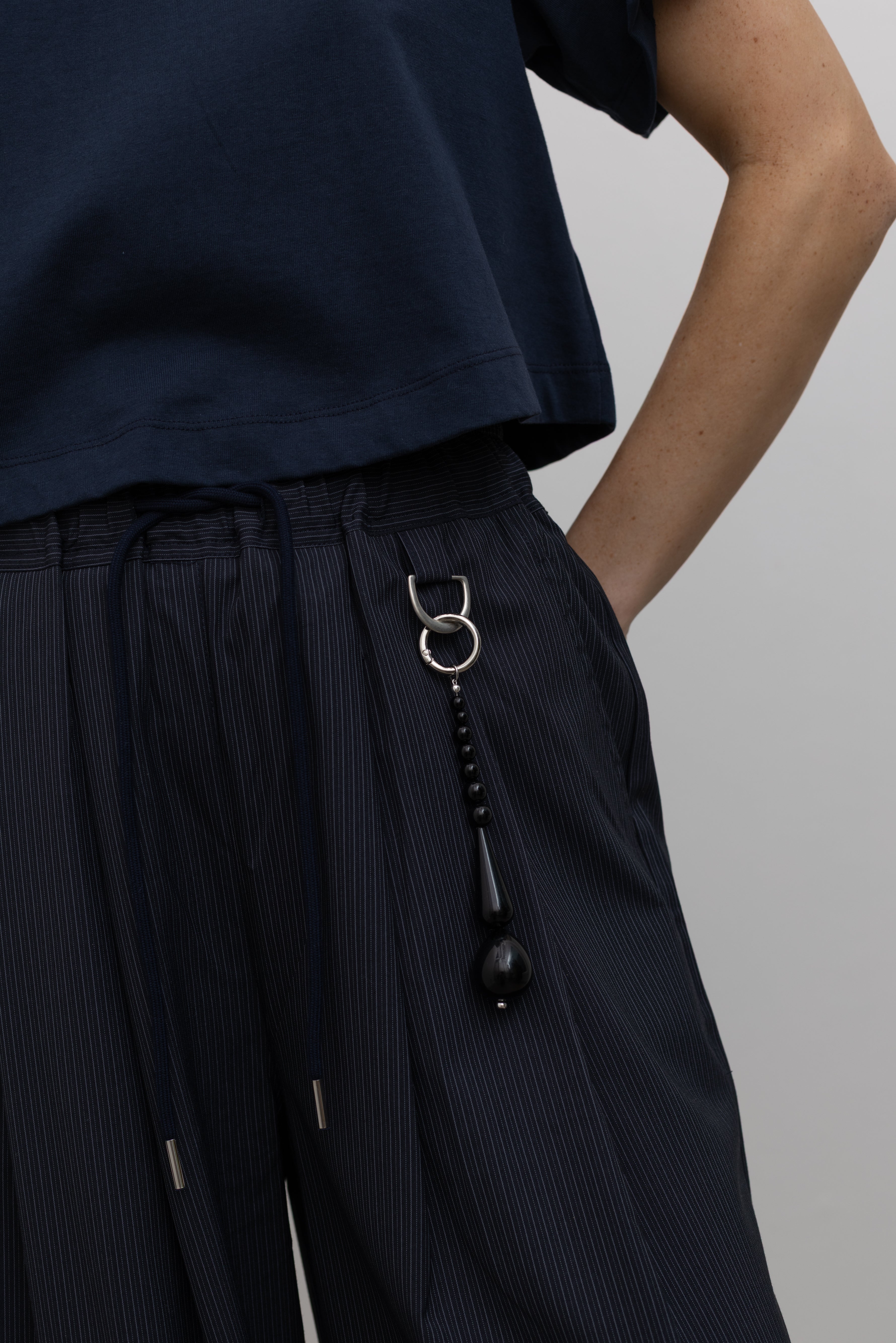 Detailed product view of Woman standing wearing Spring fashion in a pair of Collective Navy Pinstripe Wide Leg Drawstring Pants and navy blue tee with black beaded accessory at belt loop at Alas Eius