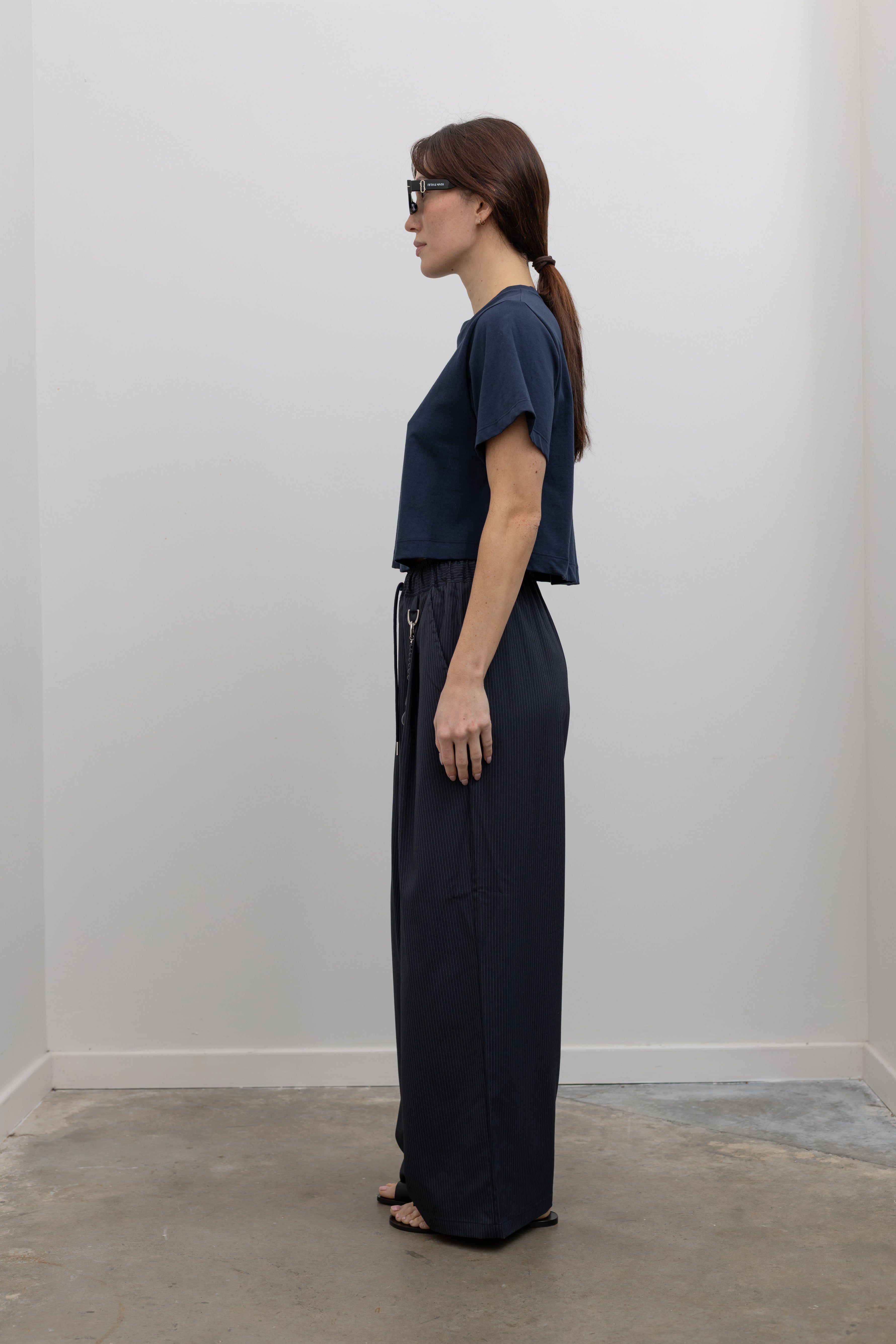 Side view of Woman standing wearing Spring fashion in a pair of Collective Navy Pinstripe Wide Leg Drawstring Pants and navy blue tee with black leather sandals at Alas Eius