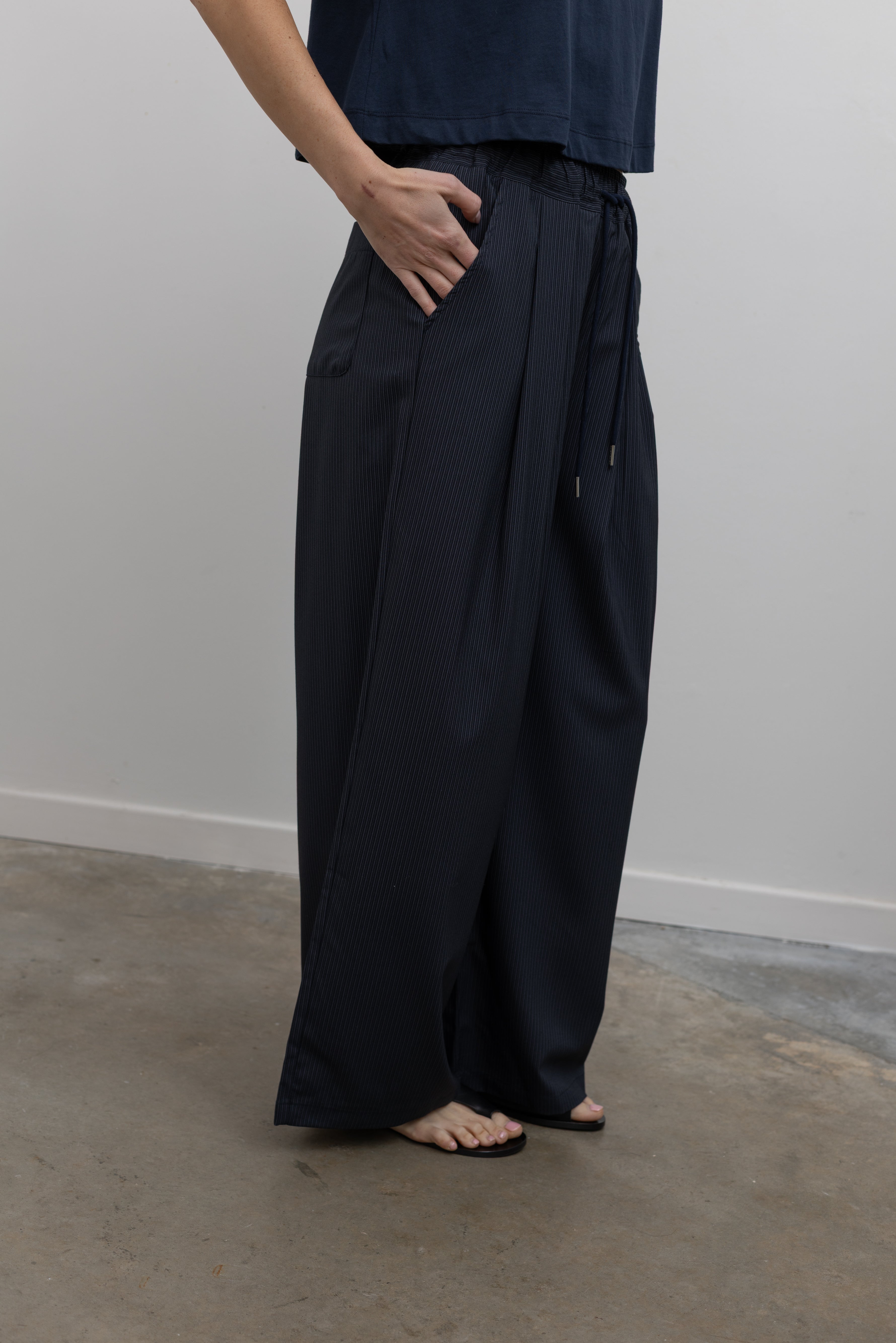 Detailed product view of Woman standing wearing Spring fashion in a pair of Collective Navy Pinstripe Wide Leg Drawstring Pants and navy blue tee with black leather sandals at Alas Eius