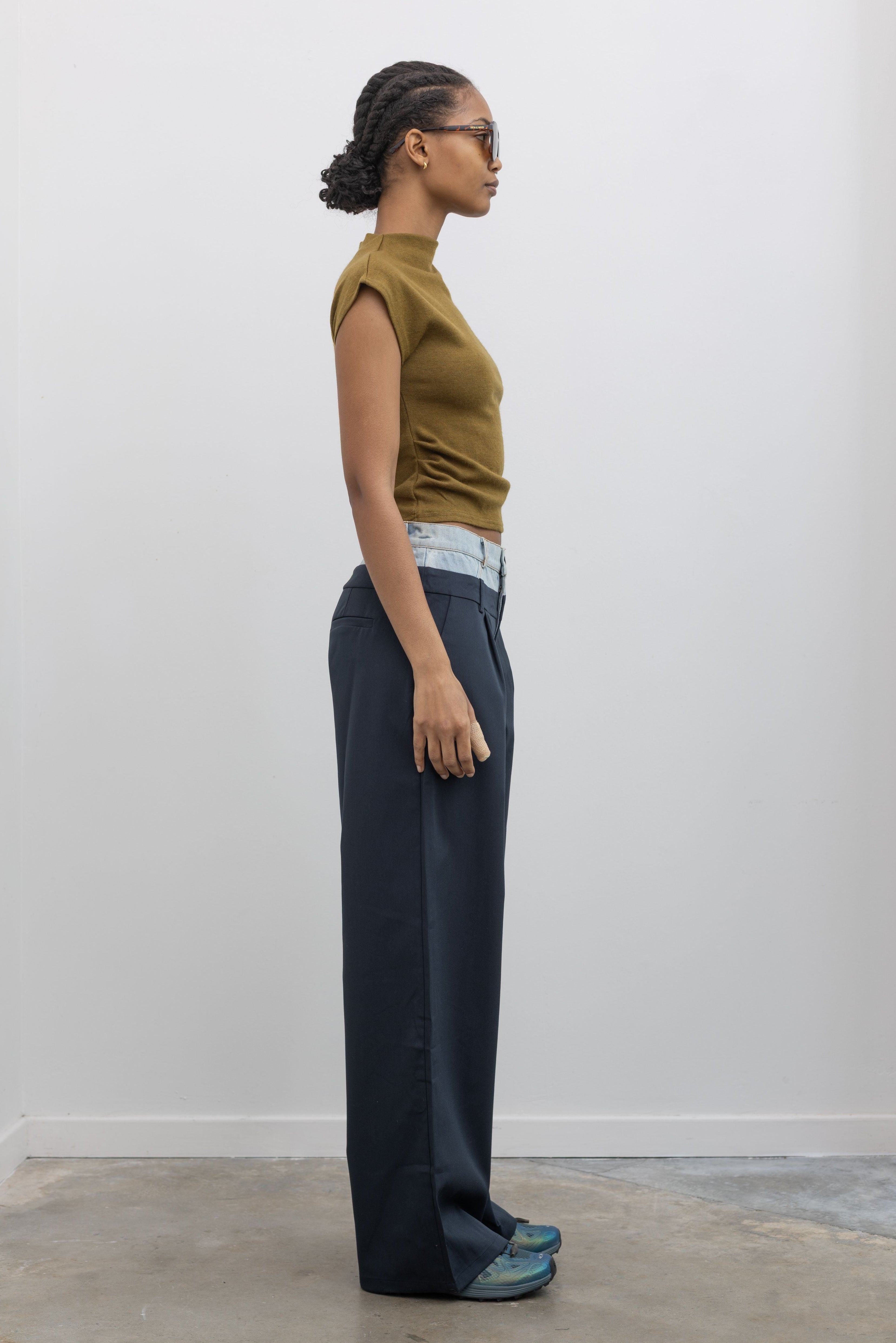 Full length front view of a woman wearing spring fashion in Collective Navy Blue pleated trouser featuring a layered look with a light wash denim waistband paired with a short sleeve mock neck top and Salomon sneakers at Alas Eius. 