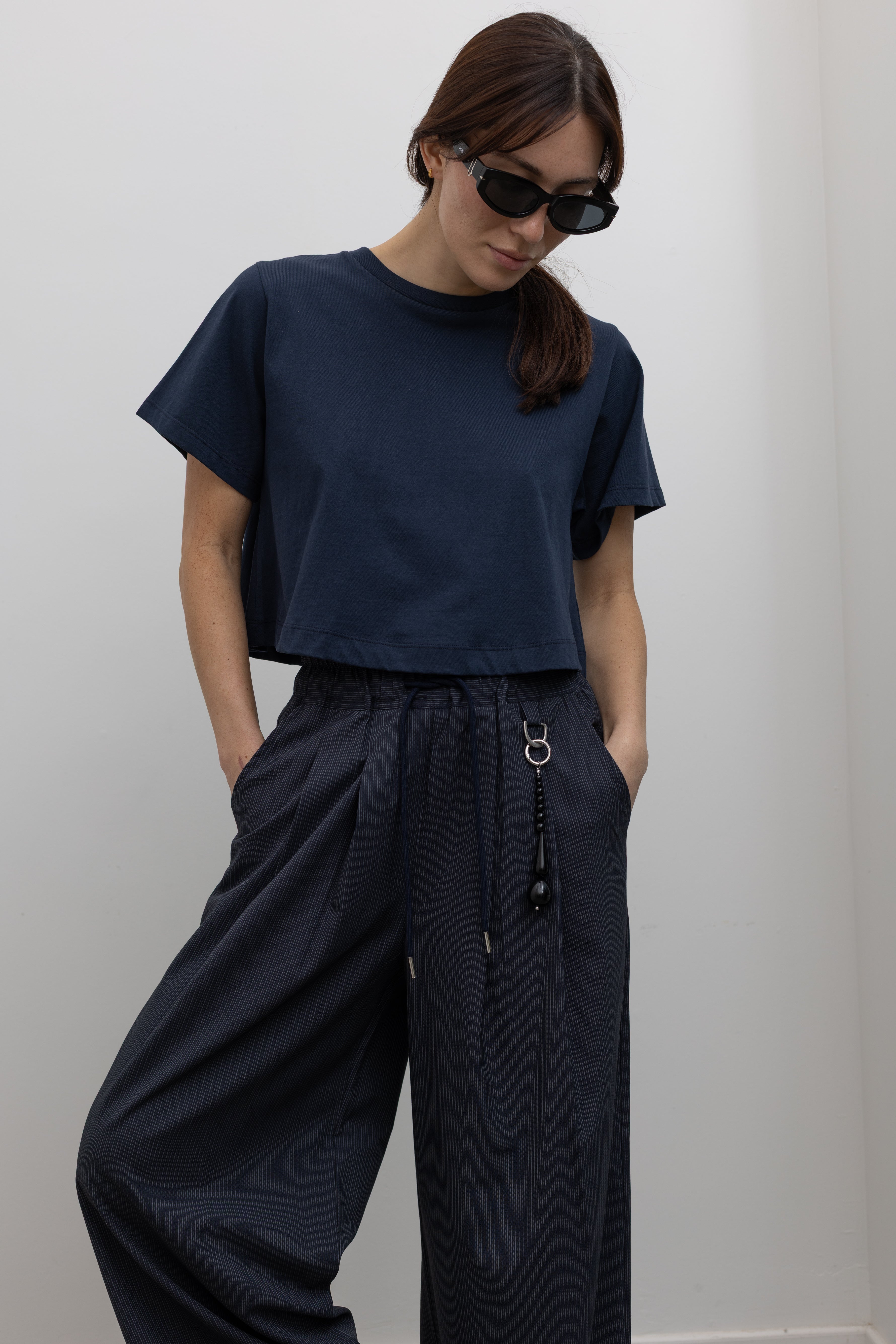 Detailed product view of Woman standing wearing Spring fashion in a pair of Collective Navy Pinstripe Wide Leg Drawstring Pants and navy blue tee with black beaded accessory at belt loop and black sunglasses at Alas Eius