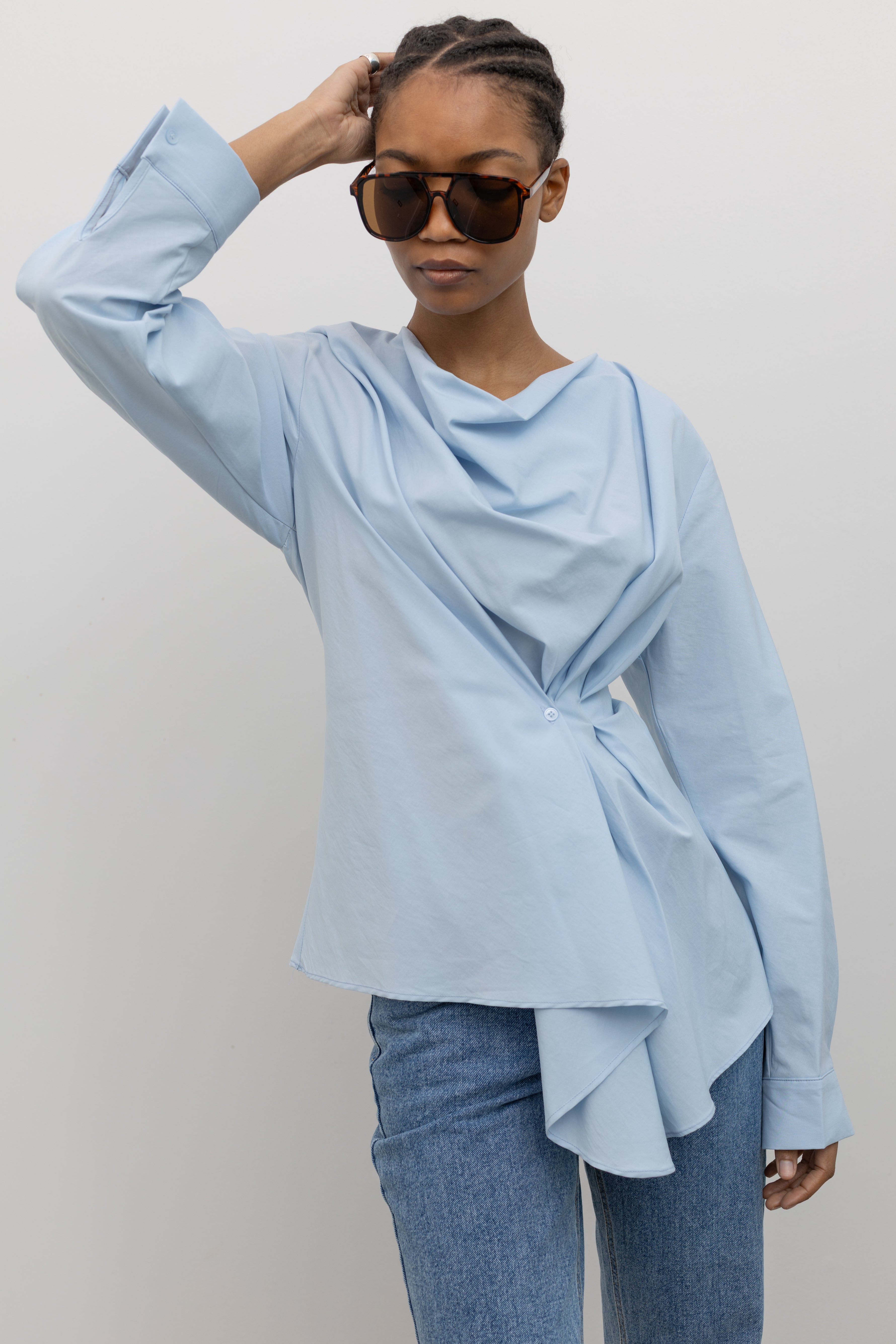Woman wearing chic and architectural spring fashion in a Collective label sky blue cotton long sleeve top featuring a fitted natural waistline with a side draped motif creating an asymmetrical hem and cowl neckline paired with light wash denim jeans at Alas Eius. 