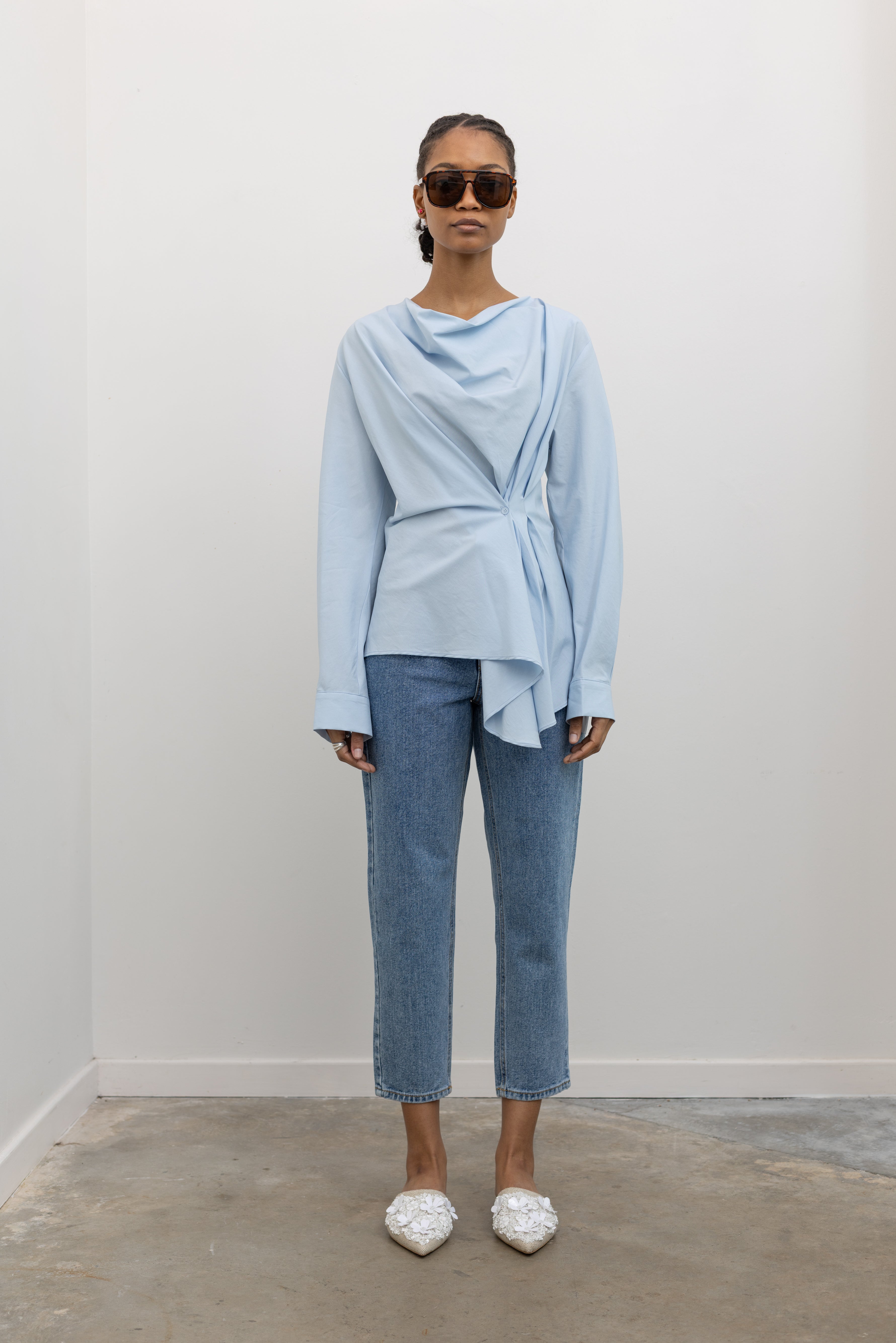 Full length front view of a woman wearing chic and architectural spring fashion in a Collective label sky blue cotton long sleeve top featuring a fitted natural waistline with a side draped motif creating an asymmetrical hem and cowl neckline paired with light wash denim jeans at Alas Eius. 
