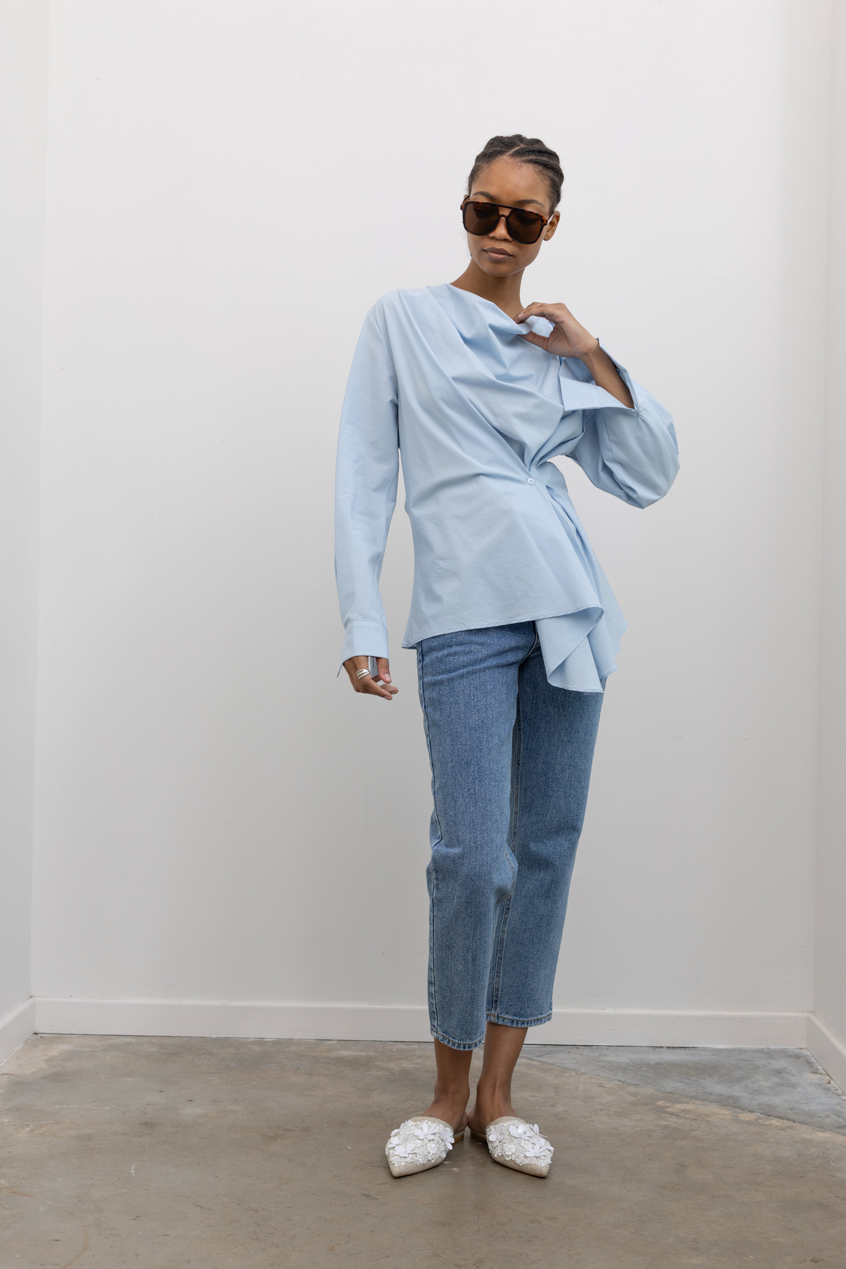 Full length front view of a woman wearing chic and architectural spring fashion in a Collective label sky blue cotton long sleeve top featuring a fitted natural waistline with a side draped motif creating an asymmetrical hem and cowl neckline paired with light wash denim jeans at Alas Eius. 