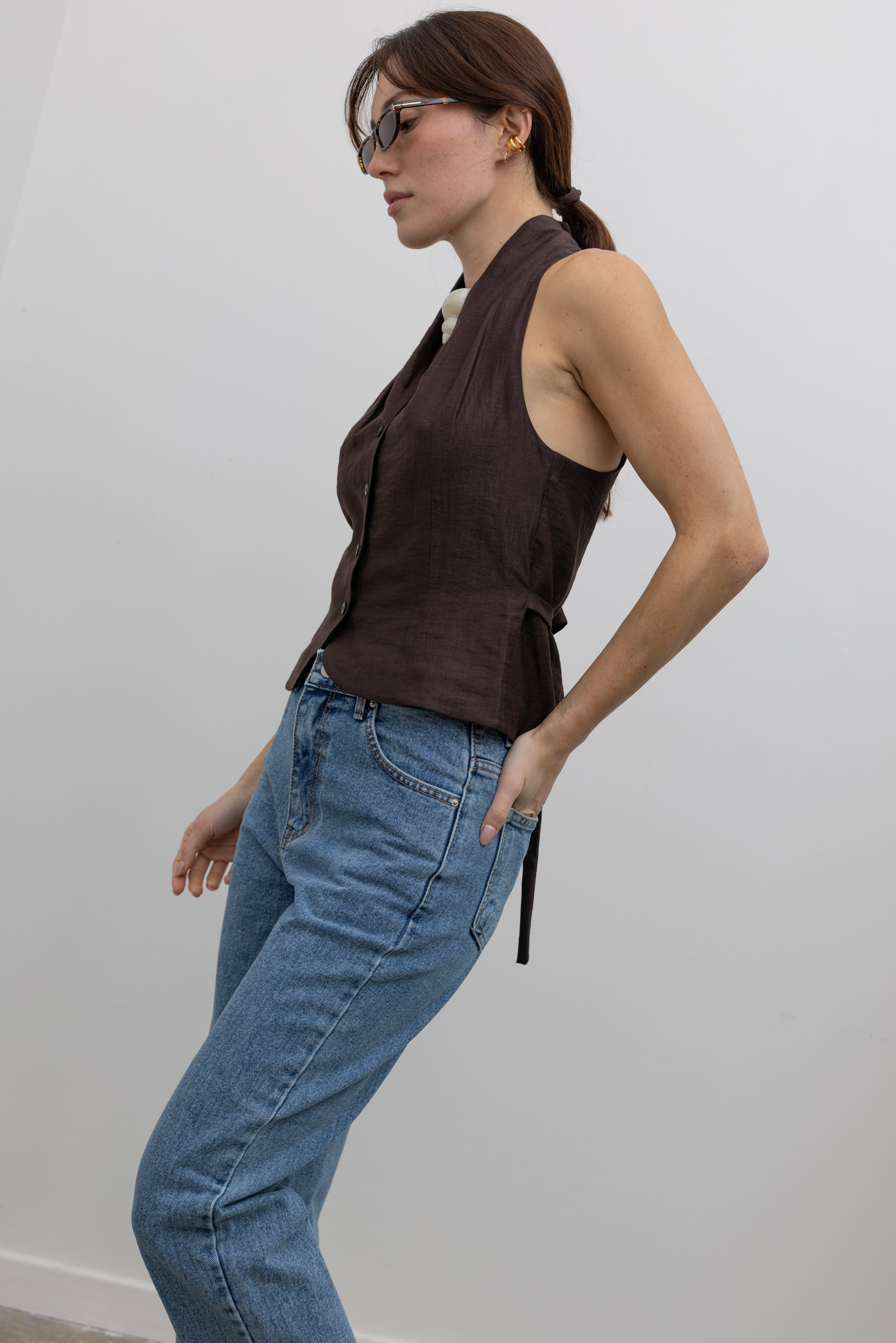 Detailed product view of Woman standing wearing Spring fashion in a brown linen pleated vest and a pair of Collective Stone Wash Carrot High Rise Jeans and sunglasses at Alas Eius