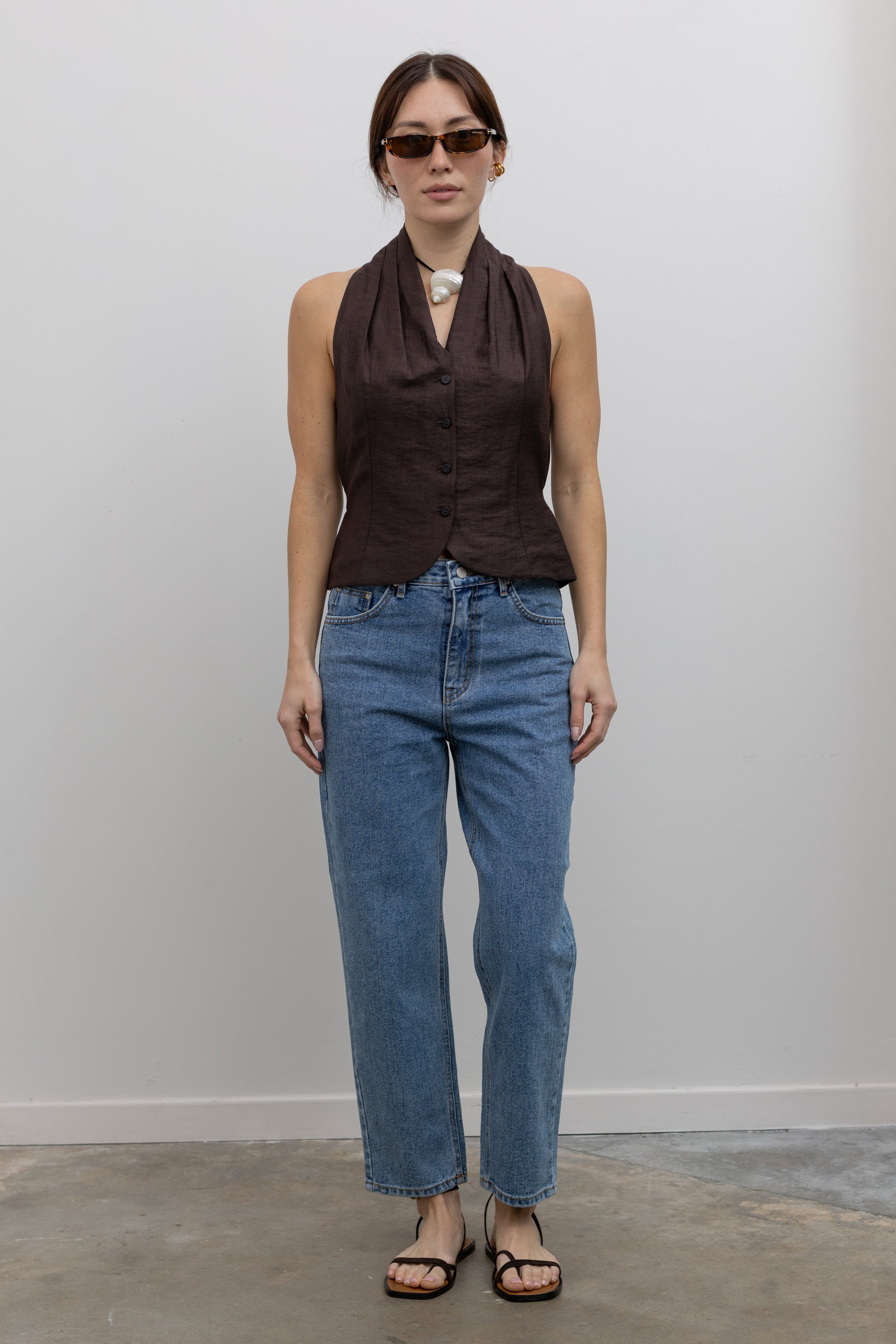 Woman standing wearing Spring fashion in a brown linen pleated vest and a pair of Collective Stone Wash Carrot High Rise Jeans with strappy sandals and sunglasses at Alas Eius