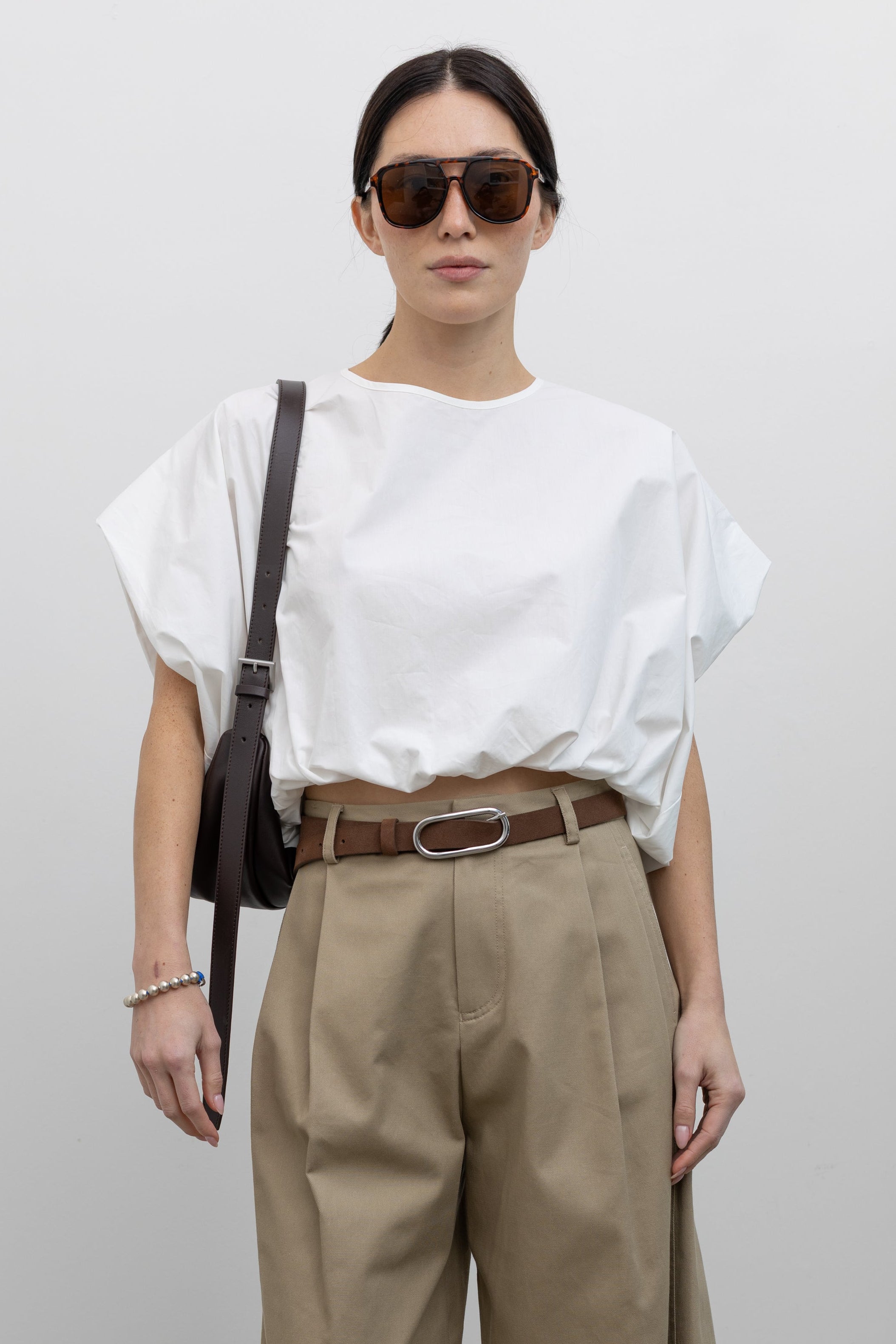 Close-up front view of a woman wearing Spring fashion in a sculptural  Collective White Cotton billow cinched top featuring draped rounded sleeves, an elasticized waistband for a fitted torso and ribbon tie at the back neck paired with timeless khaki pleated high waisted trousers and a brown leather belt at Alas Eius. 