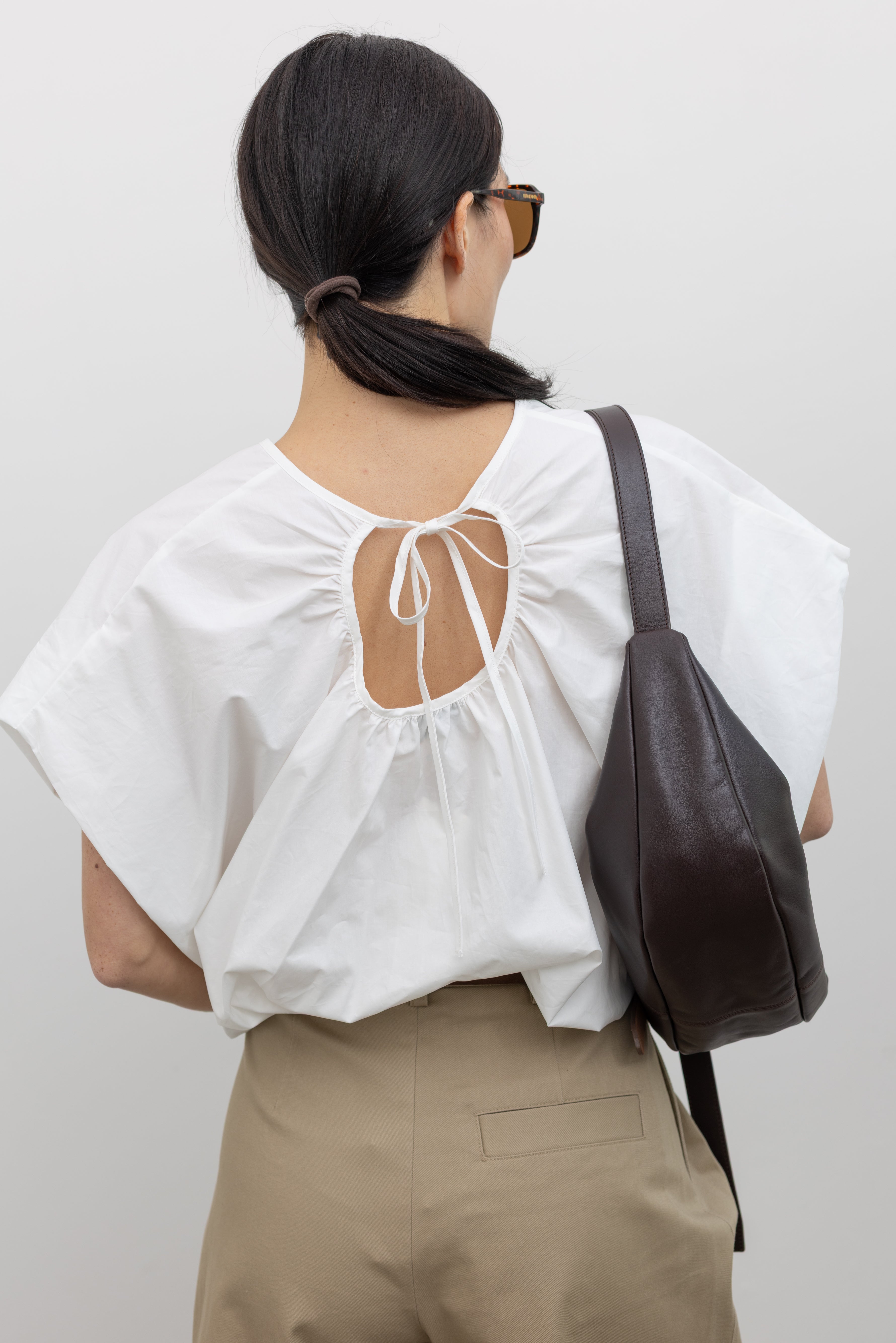 Close-up back view of a woman wearing Spring fashion in a sculptural  Collective White Cotton billow cinched top featuring draped rounded sleeves, an elasticized waistband for a fitted torso and ribbon tie at the back neck paired with timeless khaki pleated high waisted trousers and a brown leather belt at Alas Eius. 