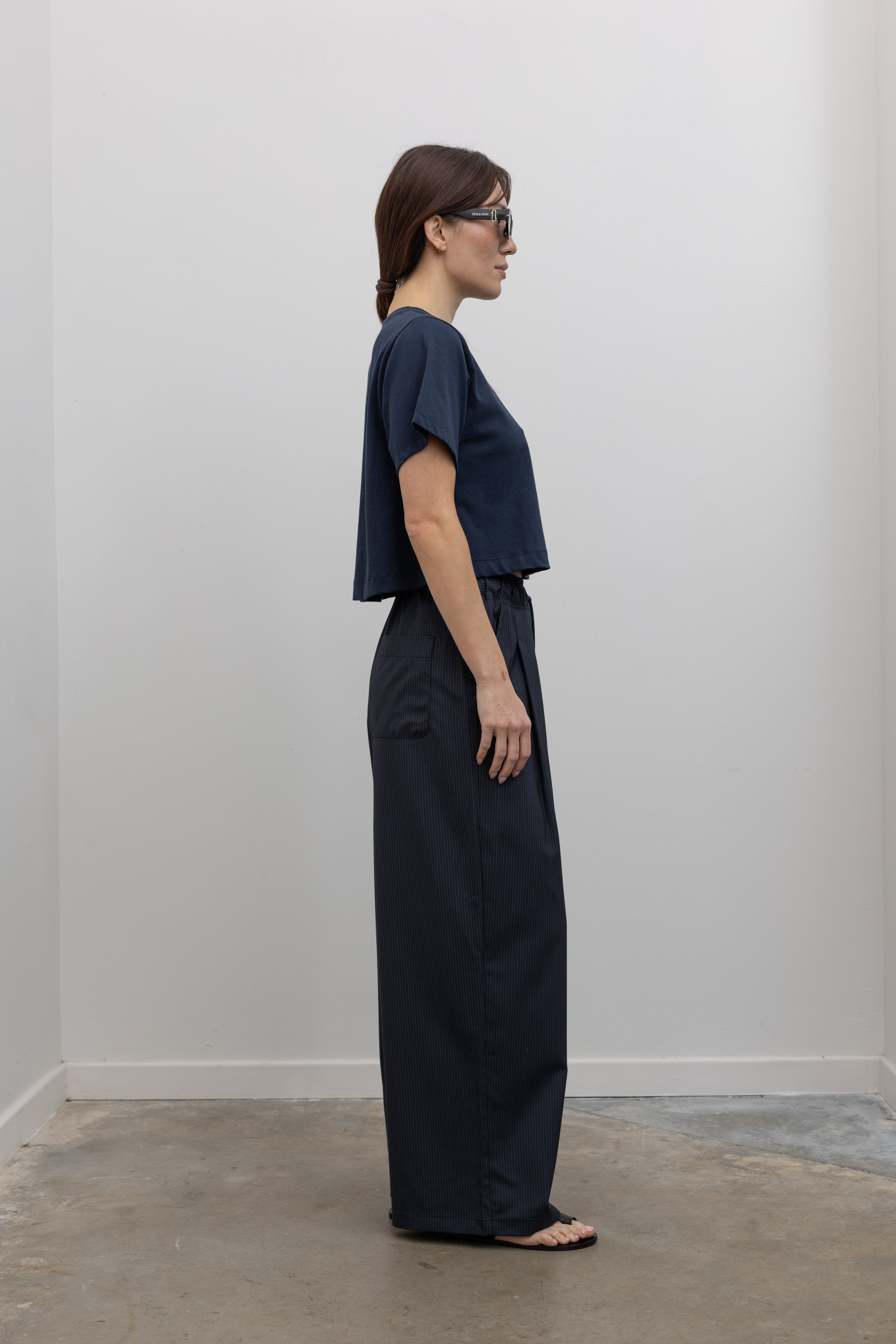 Side view of Woman standing wearing Spring fashion in a pair of Collective Navy Pinstripe Wide Leg Drawstring Pants and navy blue tee with black leather sandals at Alas Eius