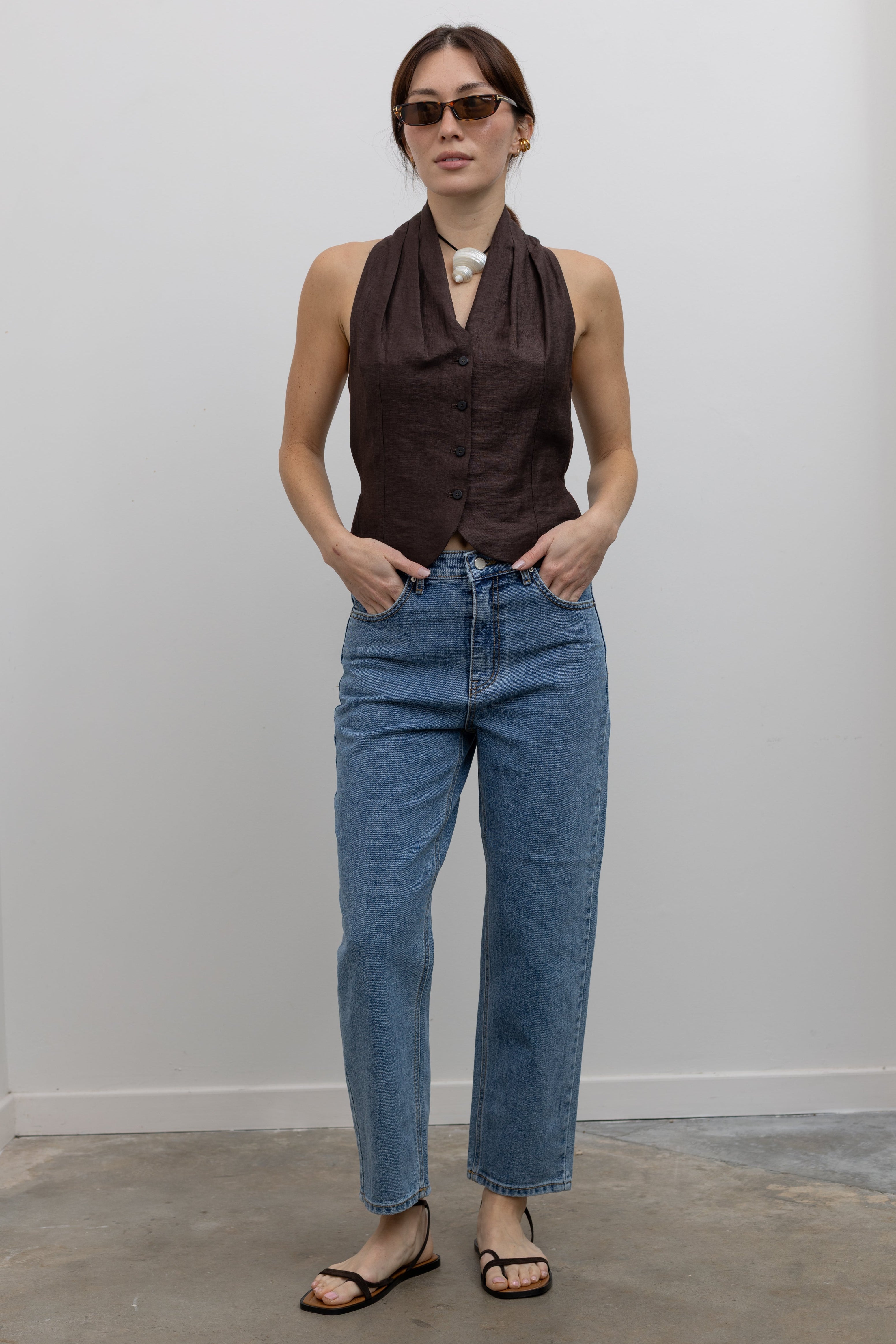 Woman standing wearing Spring fashion in a brown linen pleated vest and a pair of Collective Stone Wash Carrot High Rise Jeans with strappy sandals and sunglasses at Alas Eius