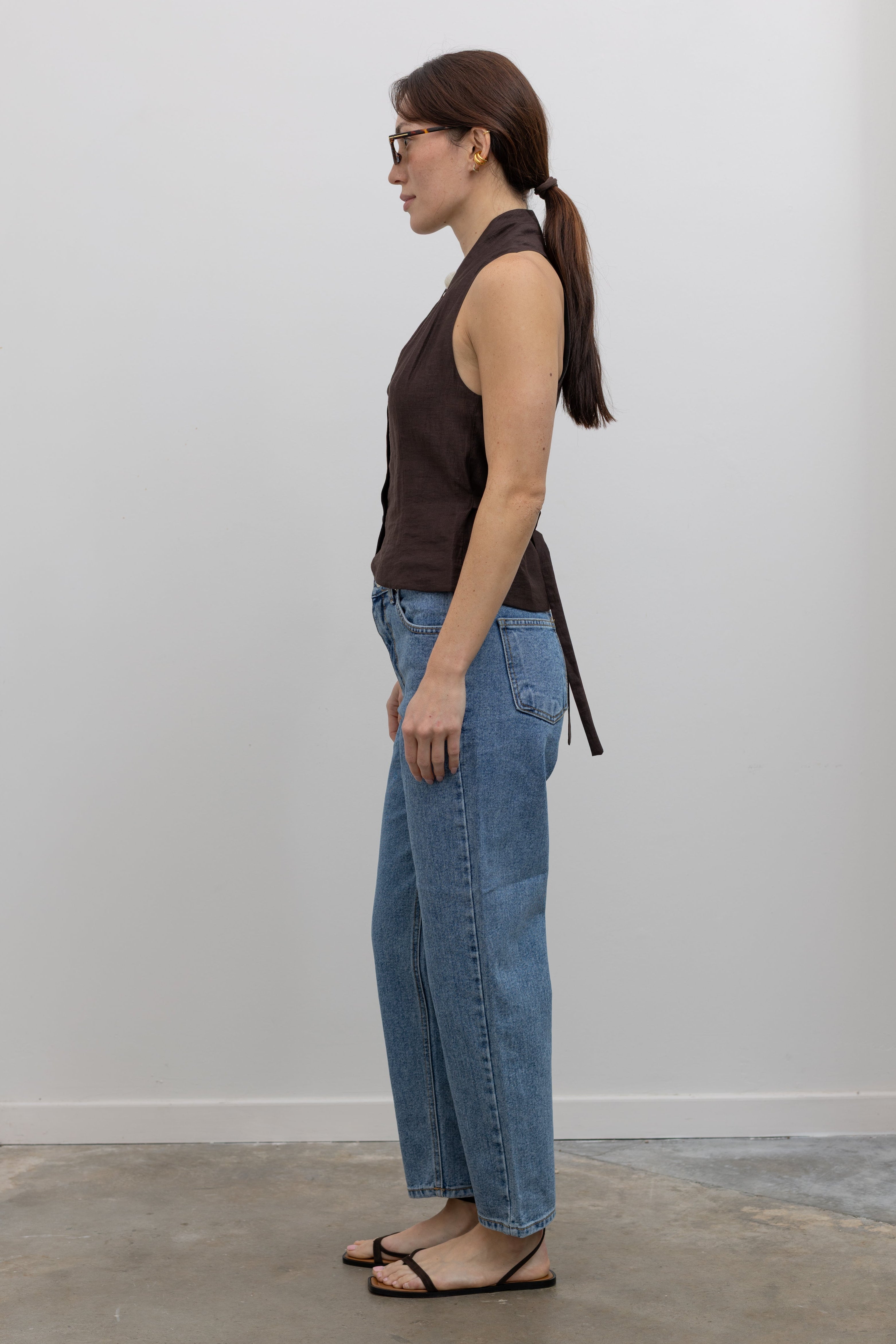 Side view of Woman standing wearing Spring fashion in a brown linen pleated vest and a pair of Collective Stone Wash Carrot High Rise Jeans with strappy sandals and sunglasses at Alas Eius