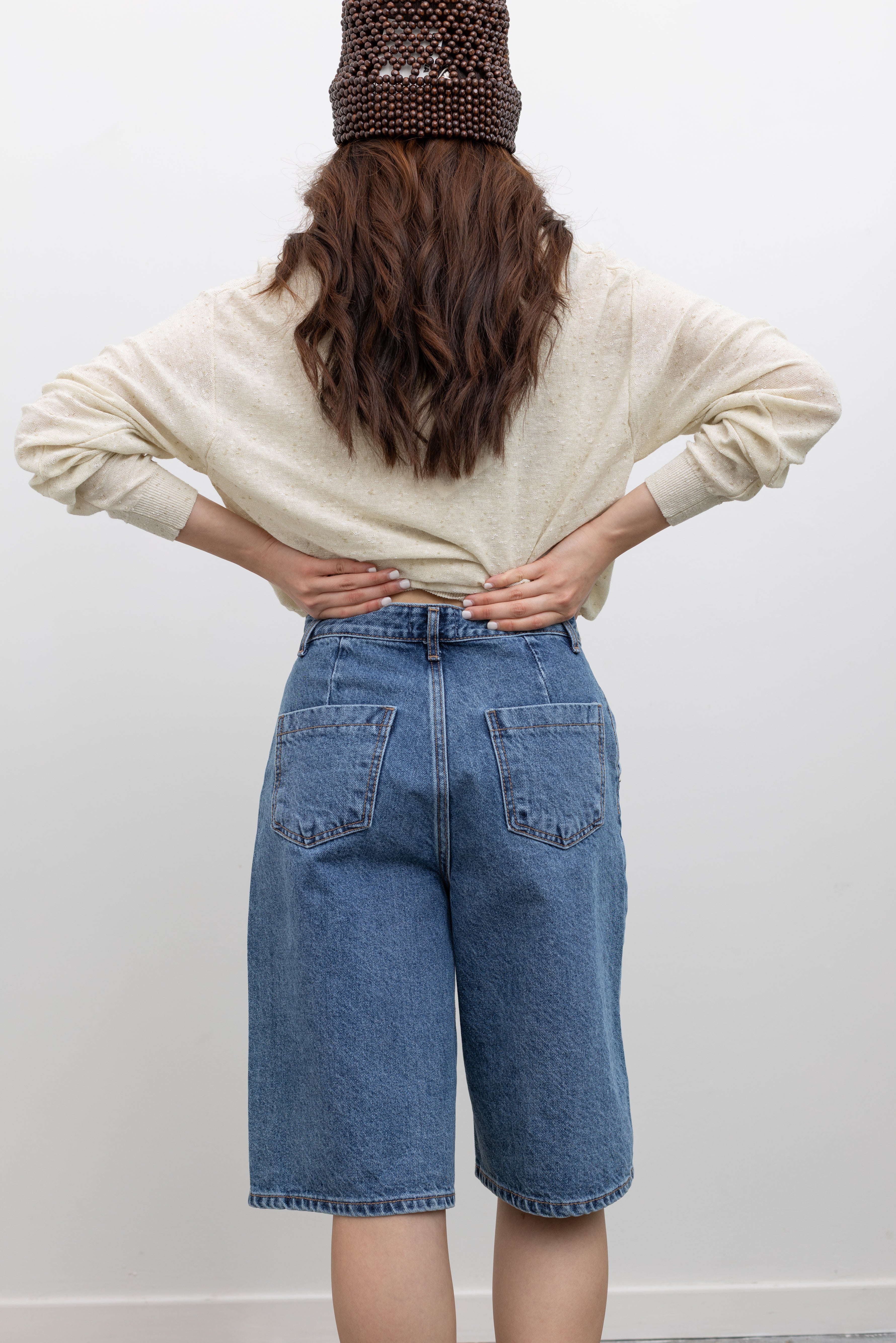 Model wearing modern summer womens fashion Collective stone-washed light blue denim bermuda shorts with a mid-to-high rise, wide-leg silhouette, and knee-length design paired with a Cordera cotton linen blend cardigan and brown wooden beaded bucket hat at Alas Eius.