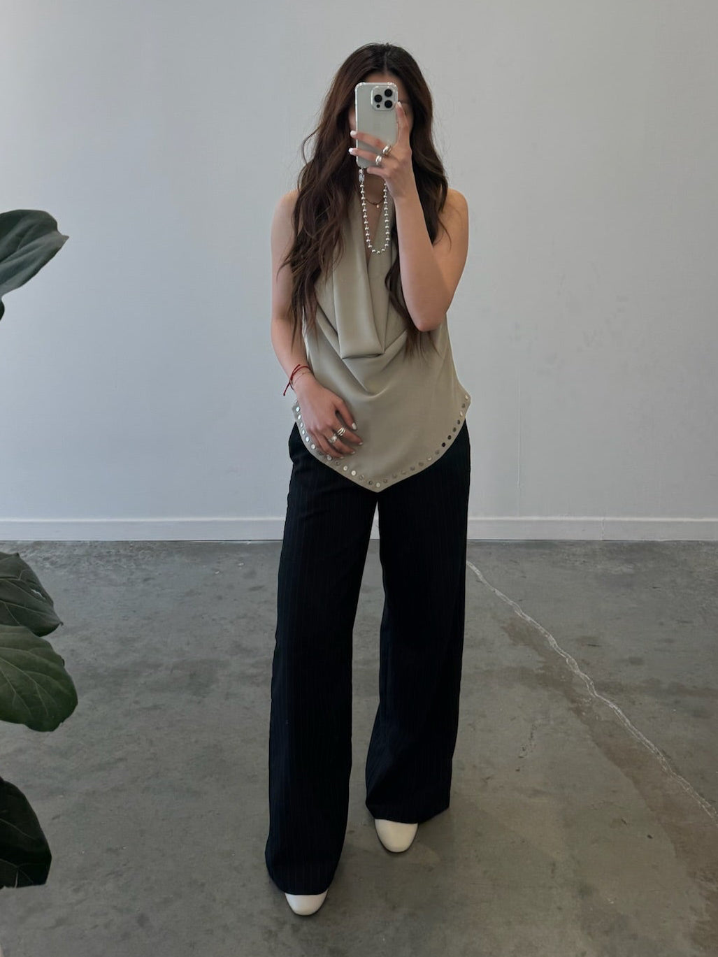 Front view of a woman wearing an edgy yet chic top in a Collective brand beige halter neck top designed with a low cowl drape, a v-cut hem that is lined with silver stud hardware, a side zipper closure, and exposed back at an Alas Eius Store.