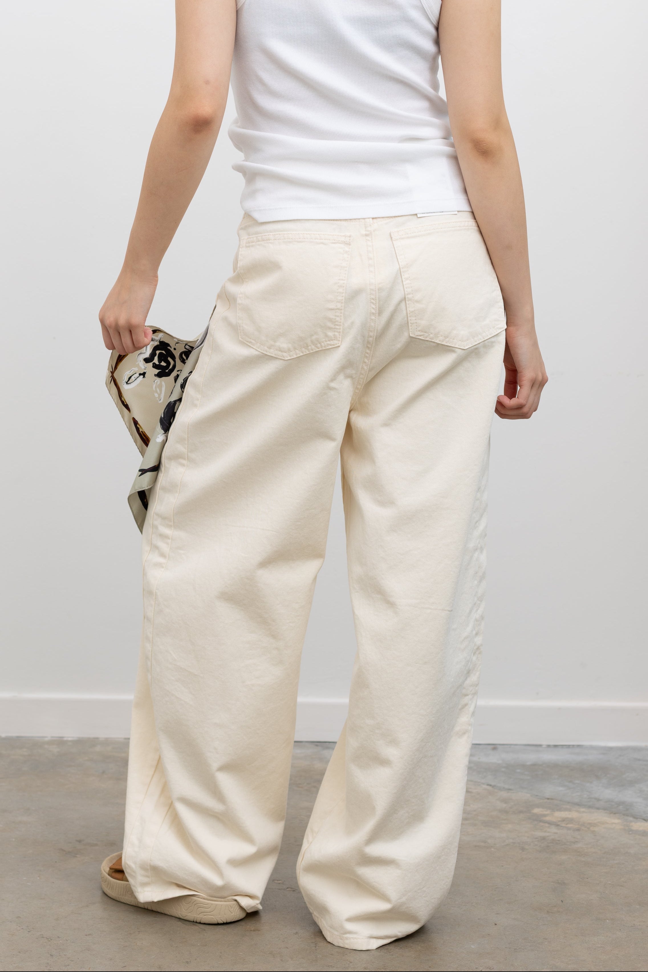Model wearing elevated summer weekend fashion in Collective ivory heavy-weight cotton denim jeans with a structured wide-leg silhouette, curved paneling, and relaxed oversized fit paired with an Alas Eius label logo tank and Veja slide sandals at Alas Eius.