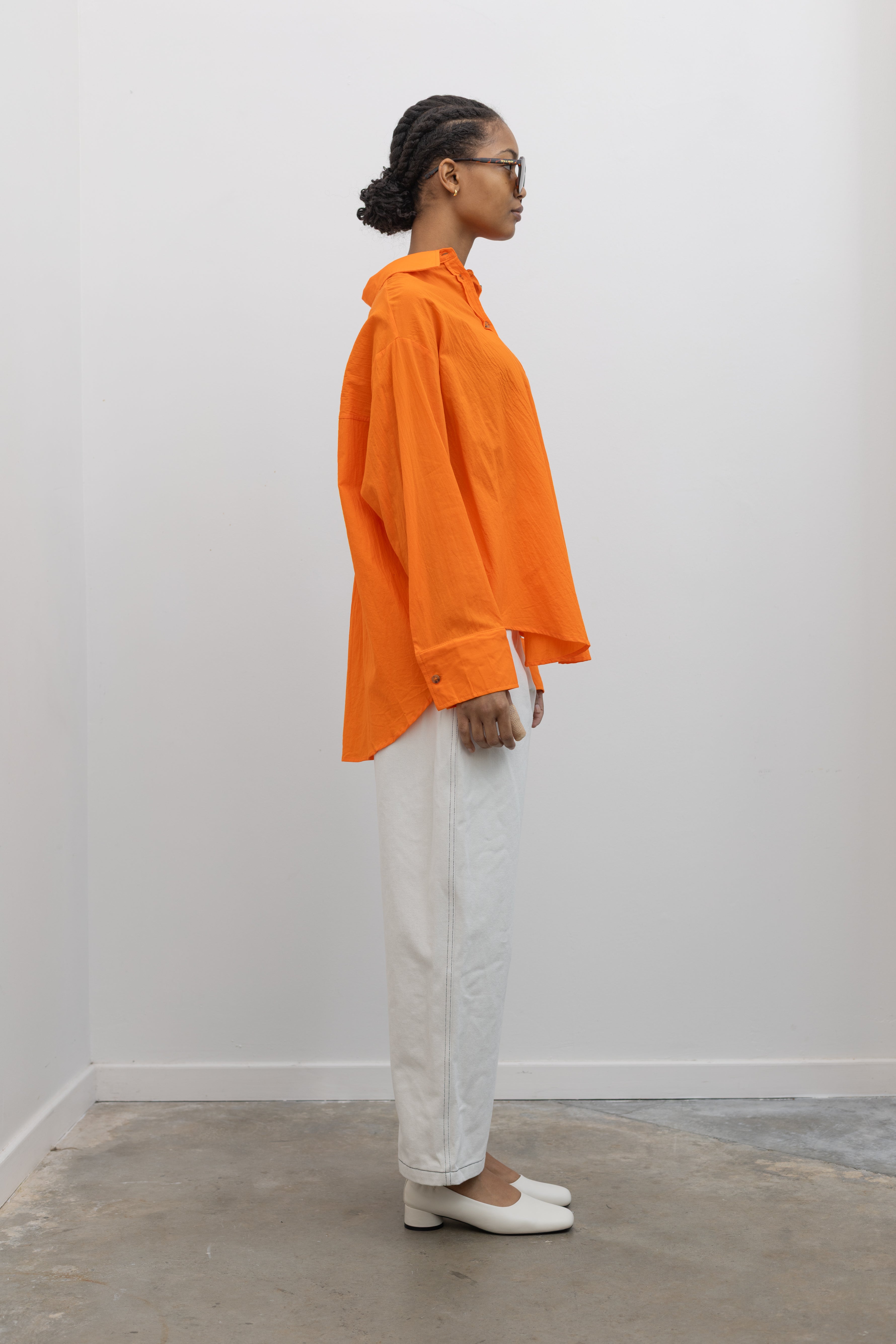 Side view of a woman wearing Spring/Summer fashion in a Collective tangerine orange cotton oversized button down collared shirt paired with white denim and sunglasses at Alas Eius.