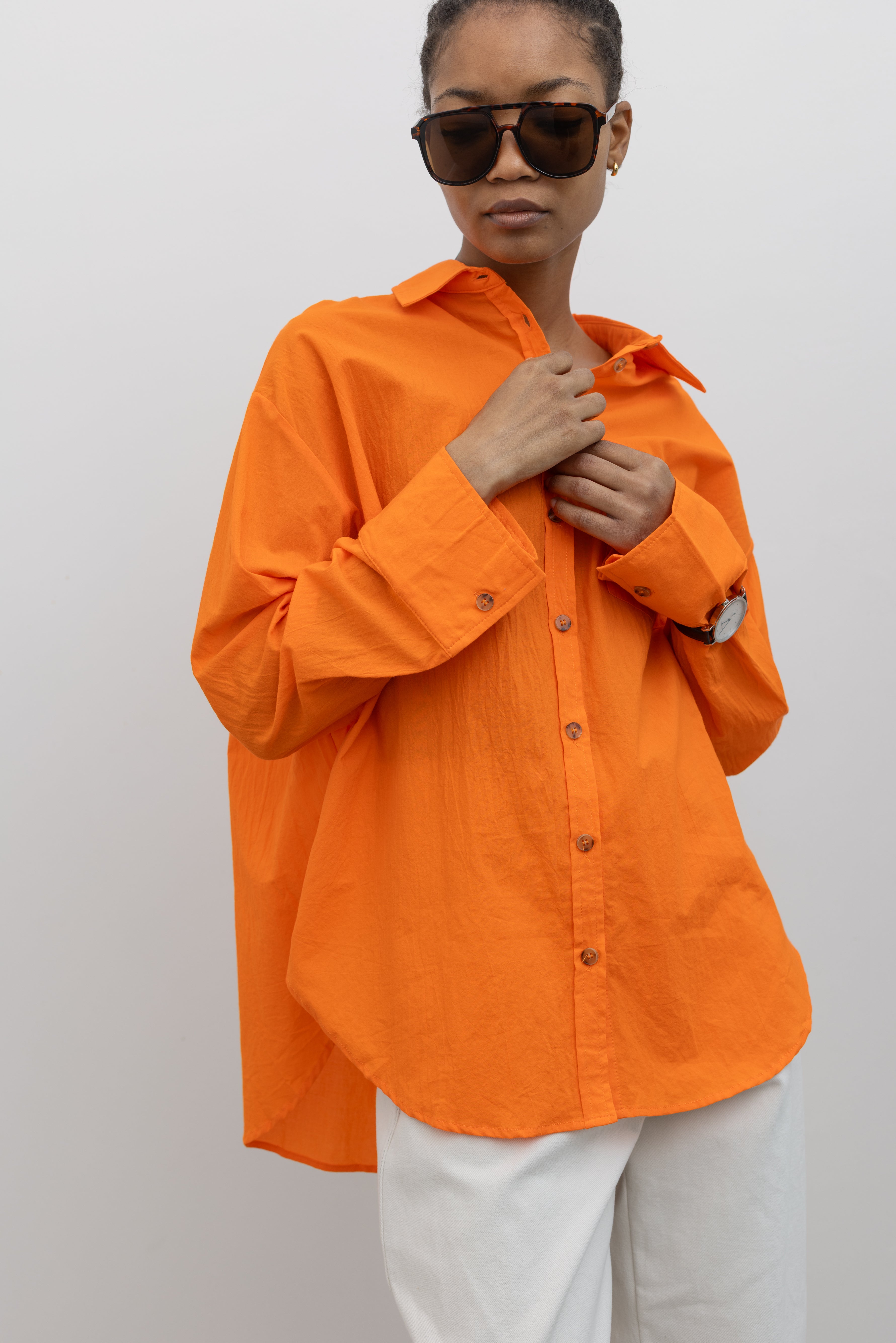Close-up product view of a woman wearing Spring/Summer fashion in a Collective tangerine orange cotton oversized button down collared shirt paired with white denim and sunglasses at Alas Eius