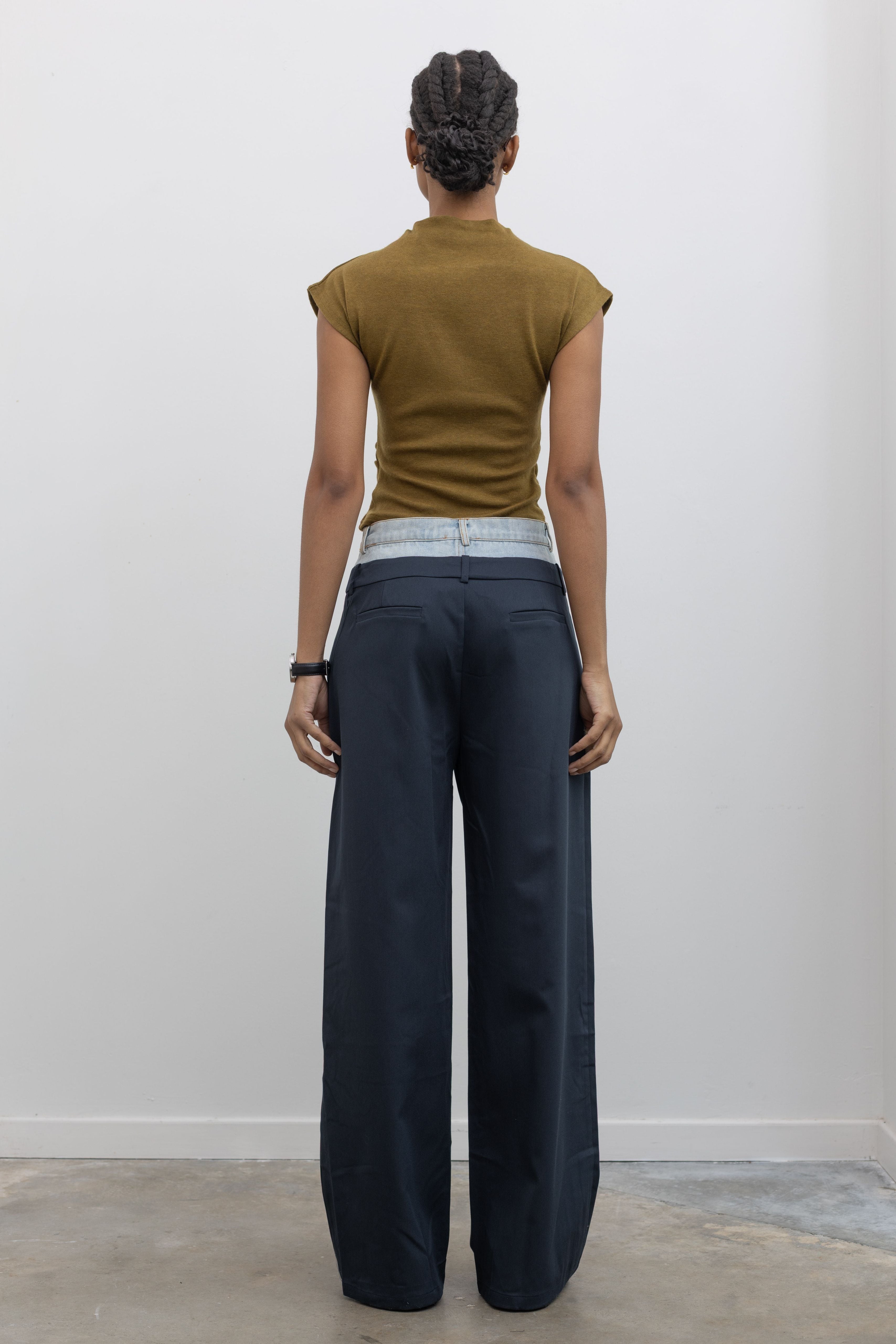 Full length back view of a woman wearing spring fashion in Collective Navy Blue pleated trouser featuring a layered look with a light wash denim waistband paired with a short sleeve mock neck top and sneakers at Alas Eius. 